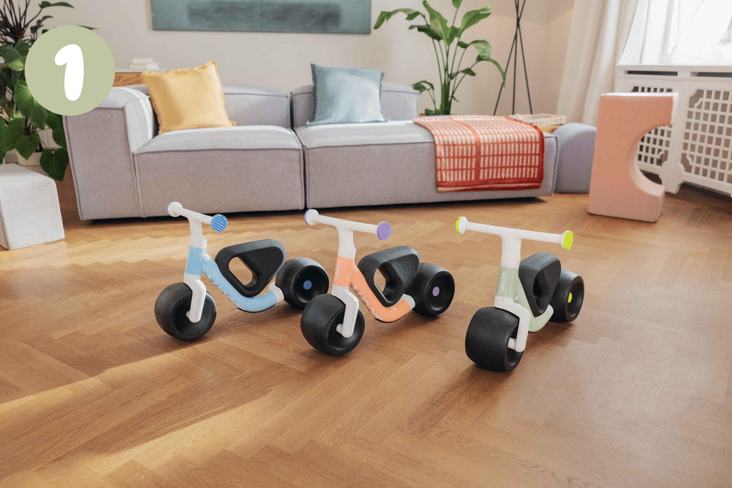 Three woom WOW self-balancing bikes in light blue, peachy orange & sage green on parquet flooring in a well-lit living room.