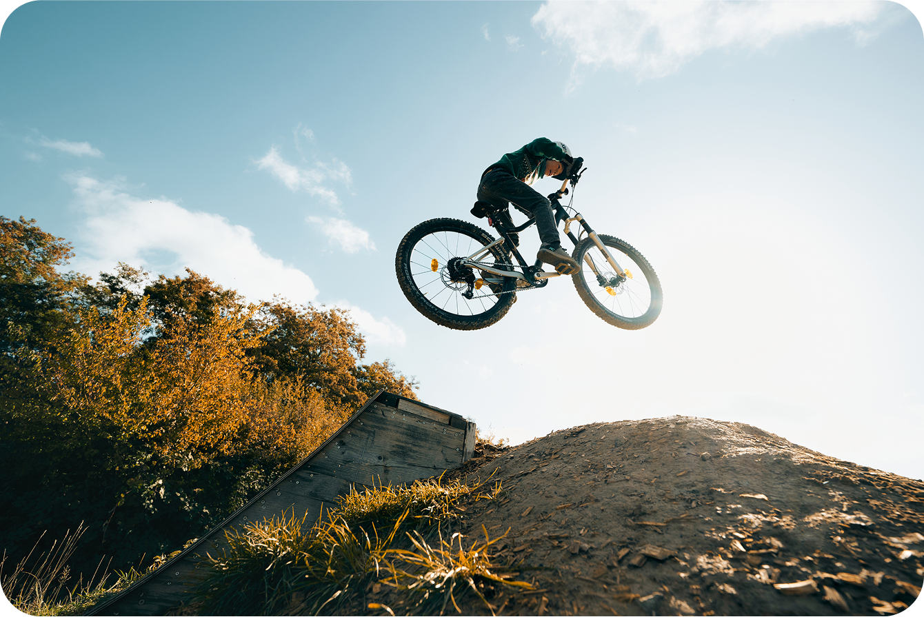 A mountain biker jumps high off a dirt ramp against a sunny sky.