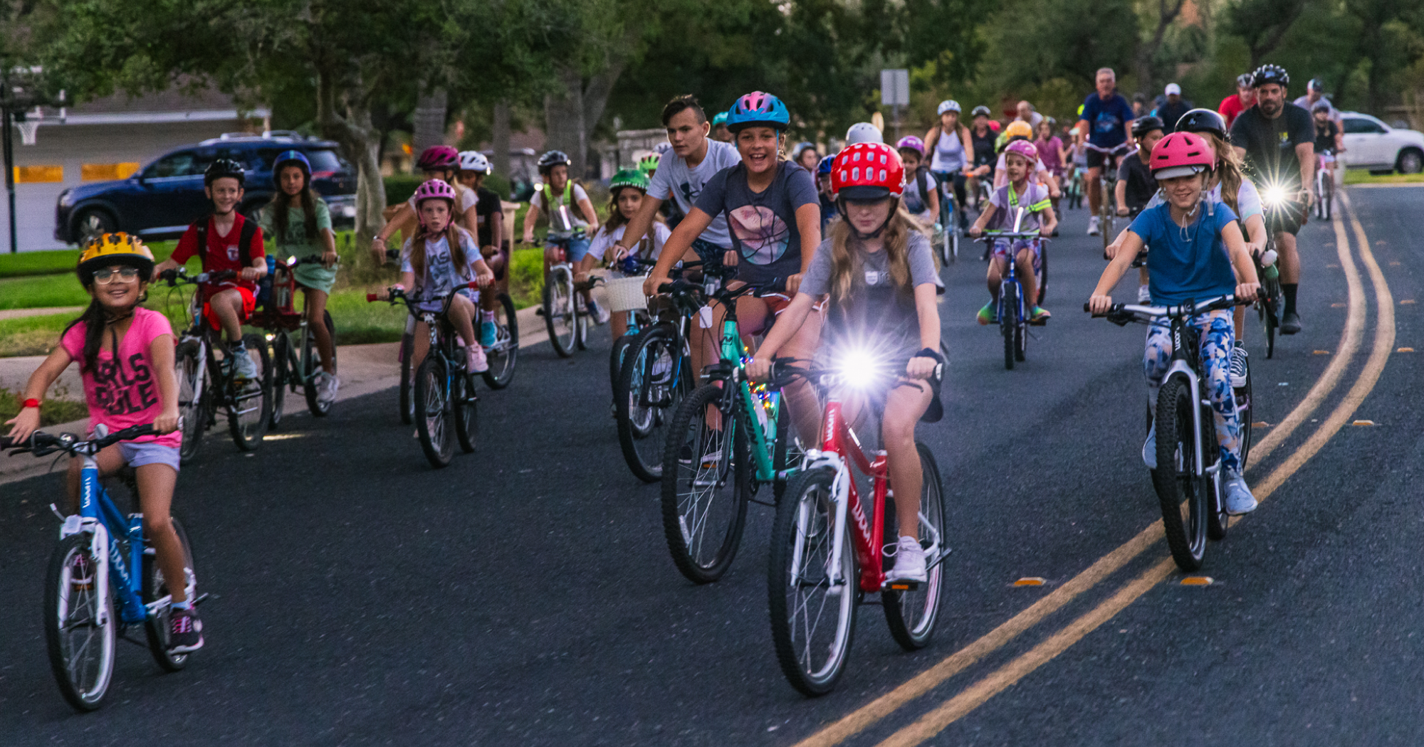 A neighborhood street is crowded by kids on bikes, with two girls on blue and red woom bikes leading the charge.