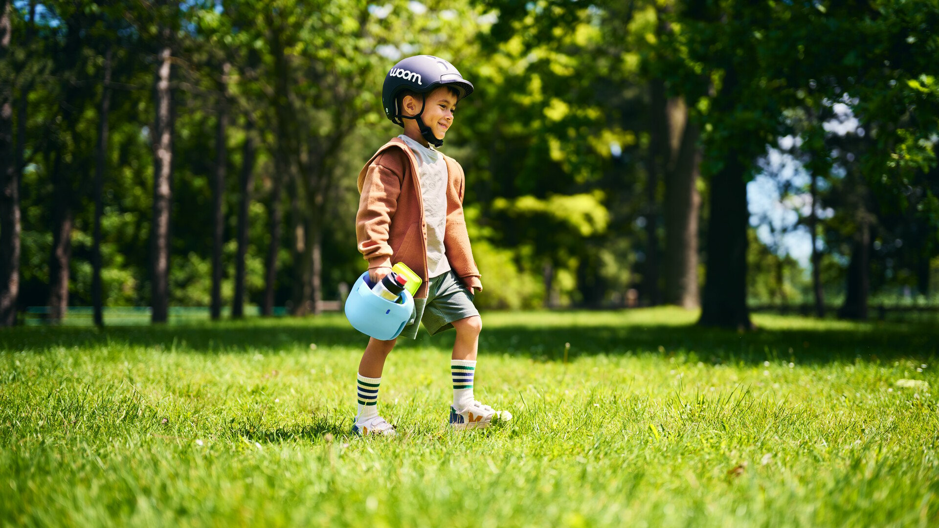 Un petit garçon avec un casque noir court dans un champ. Dans sa main droite, il tient un panier à vélo bleu ciel avec une gourde à l’intérieur.