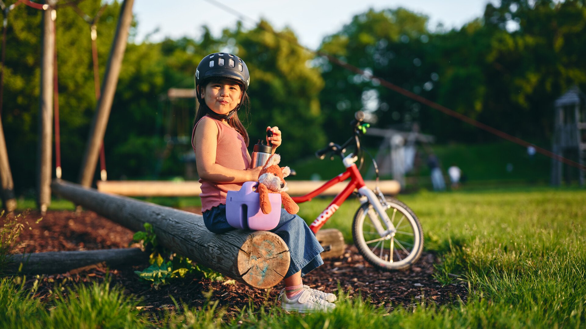 Een klein meisje met zwart haar, donkere ogen en een zwarte woom-helm op zit in een speeltuin midden in een veld. Naast haar staat een lilakleurig woom-fietsmandje waaruit een knuffelvosje steekt. In haar hand houdt ze de bidon van woom.