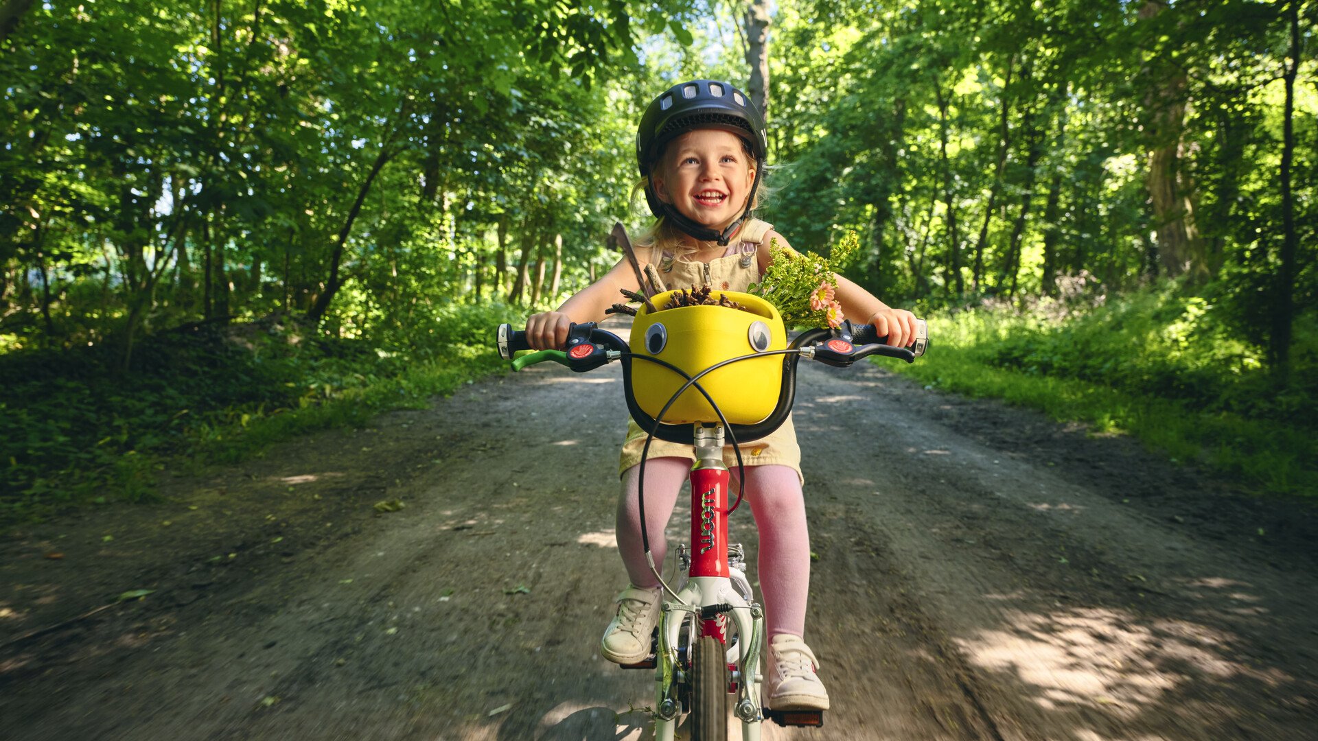 En liten, skrattande flicka med svart woom-hjälm cyklar på en rödvit woom-cykel på en skuggig skogsväg. På styret sitter en gul cykelkorg som är full med små skatter från naturen.