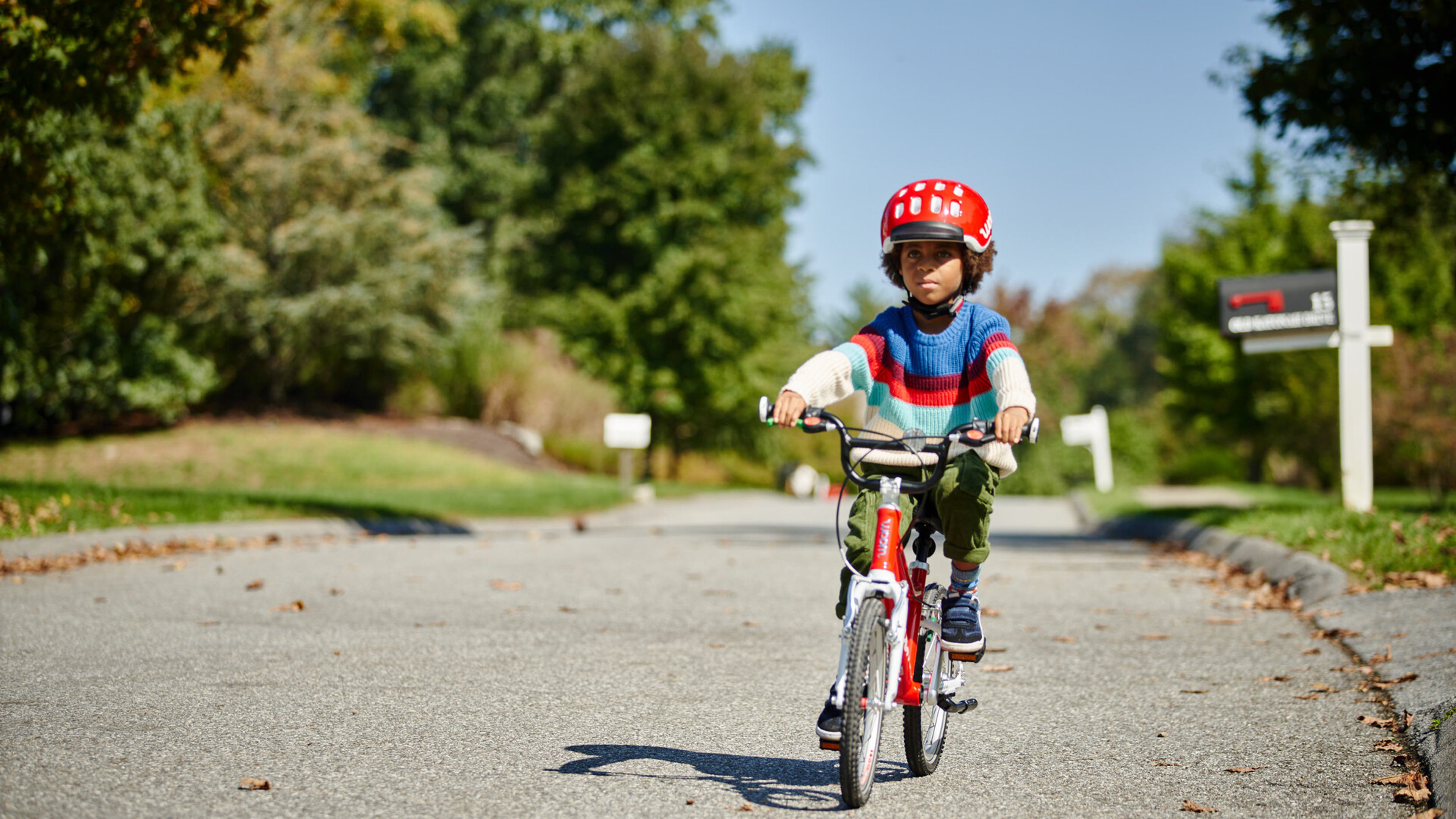 Child wearing a striped sweater and woom red KIDS' Helmet, riding a red woom ORIGINAL bike through a neighborhood.