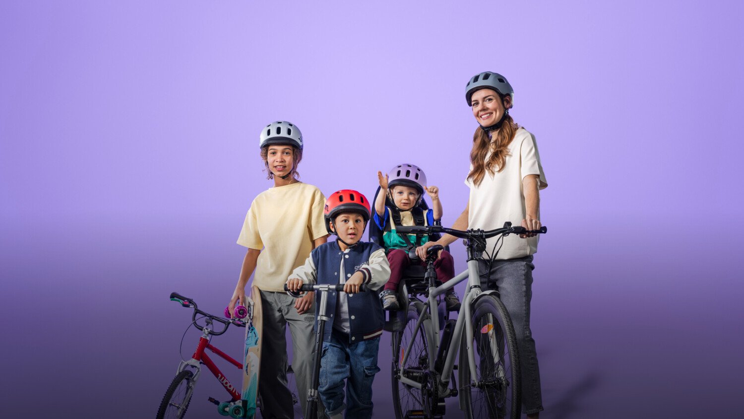 Frau mit Kleinkind im Radsitz, daneben 2 Schulkinder mit Skateboard bzw. Scooter, alle Kinder tragen den READY Helm von woom
