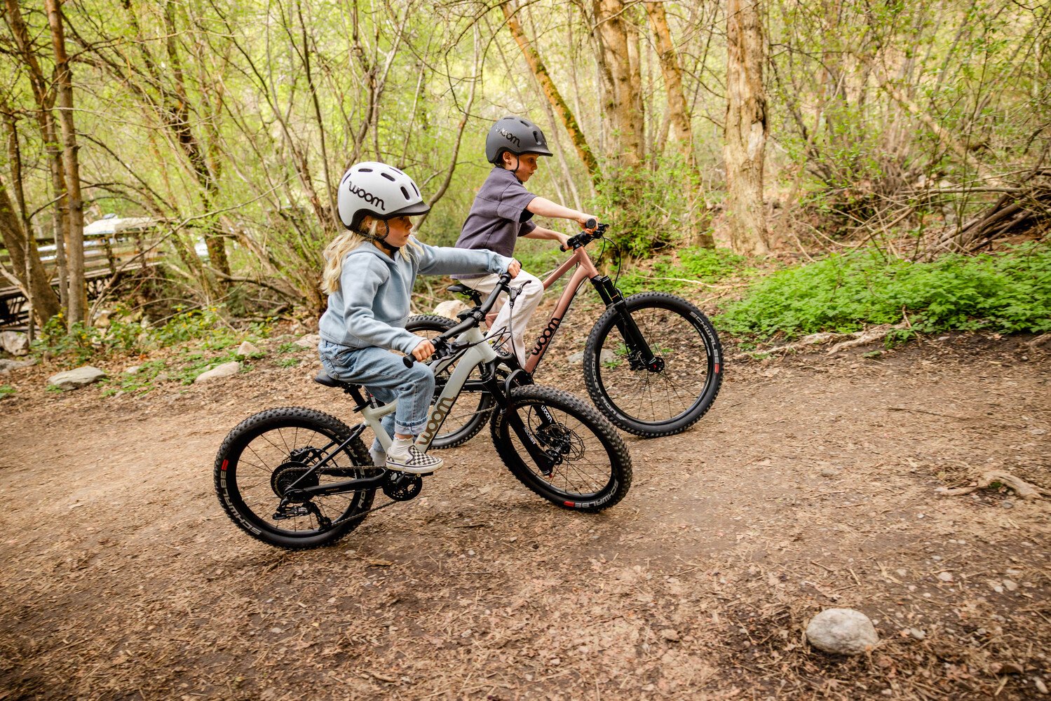 Zwei Kinder mit READY Fahrradhelmen fahren auf woom OFF AIR Kindermountainbikes im Wald über einen Trail mit Blättern.