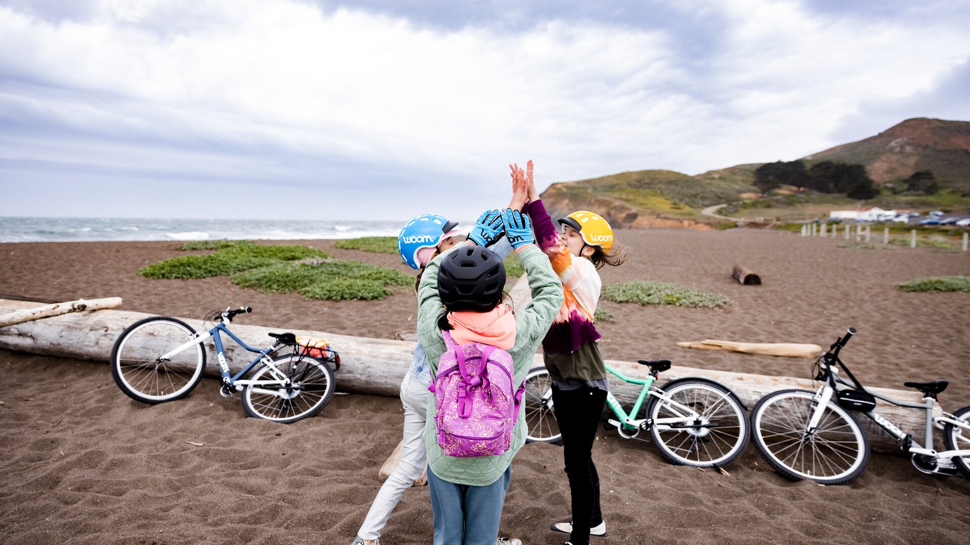 Drei Kinder, die sich am Strand ein High Five geben. Ihre Fahrräder lehnen im Hintergrund an einem Baumstamm.