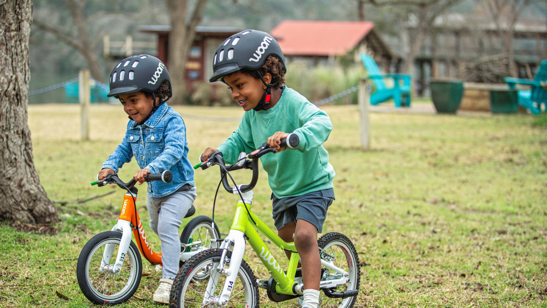 Two children in black woom helmets are riding their balance bikes across some grass.