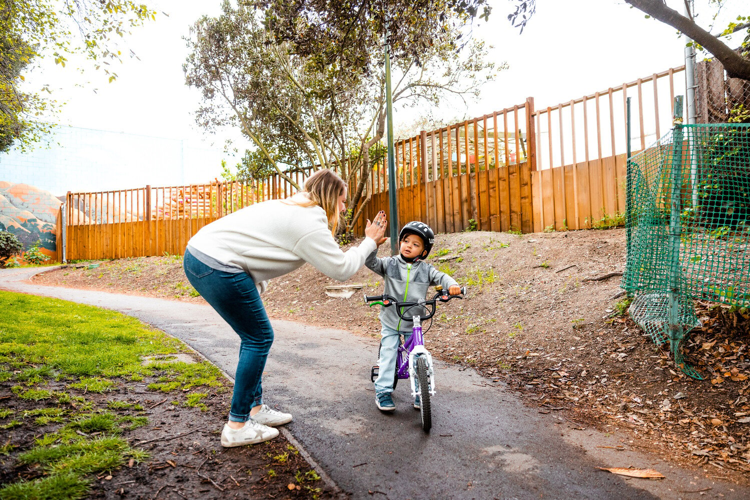 En mor og et barn klapper triumferende hinanden i hænderne. Barnet sidder på en lilla woom-børnecykel.
