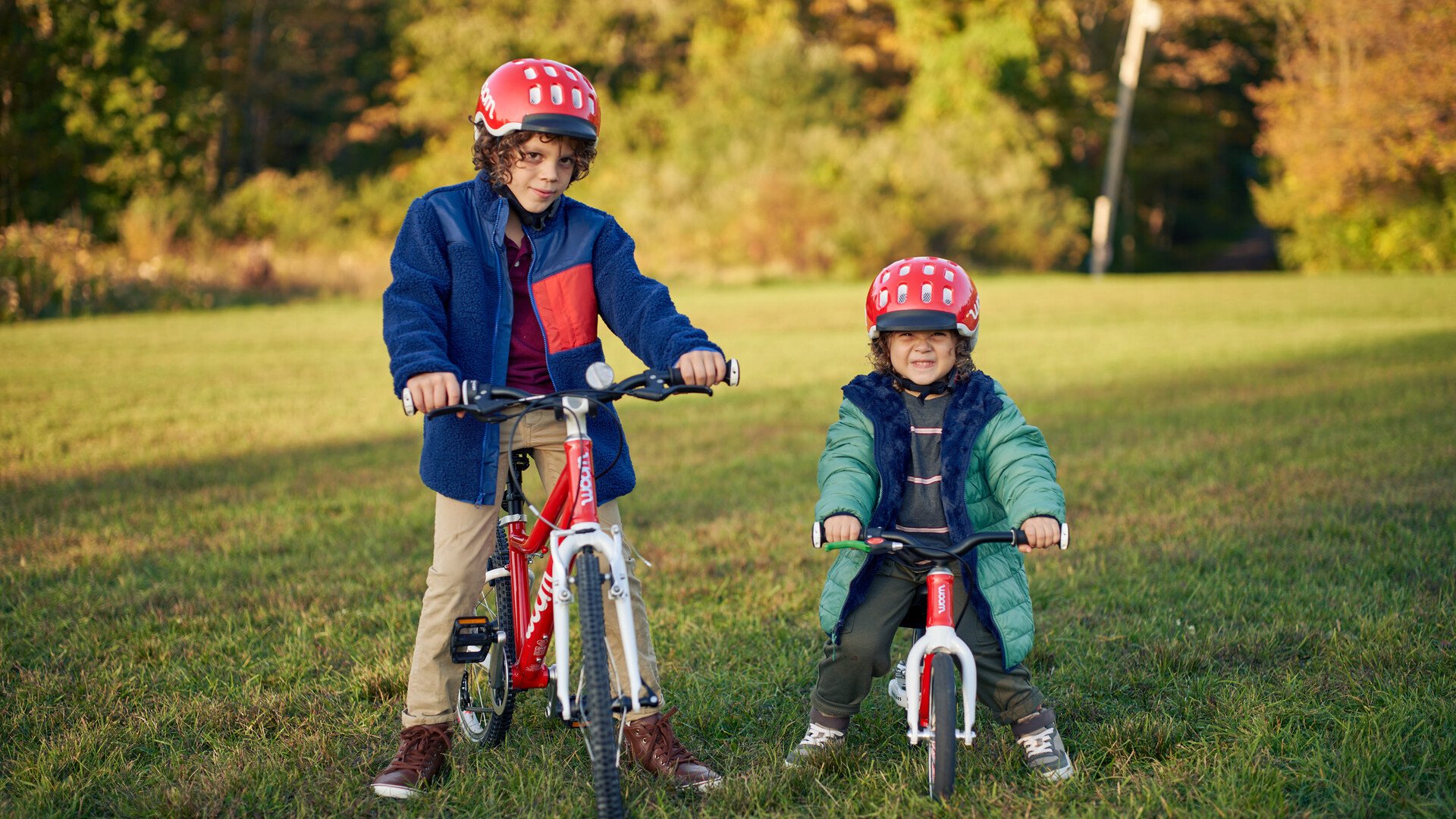 Two kids in red helmets stand with a red bike and a red balance bike. 