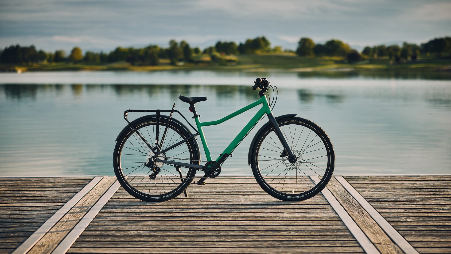 Grünes woom EXPLORE Fahrrad mit PICKUP Gepäckträger, im Hintergrund ist ein Gewässer.