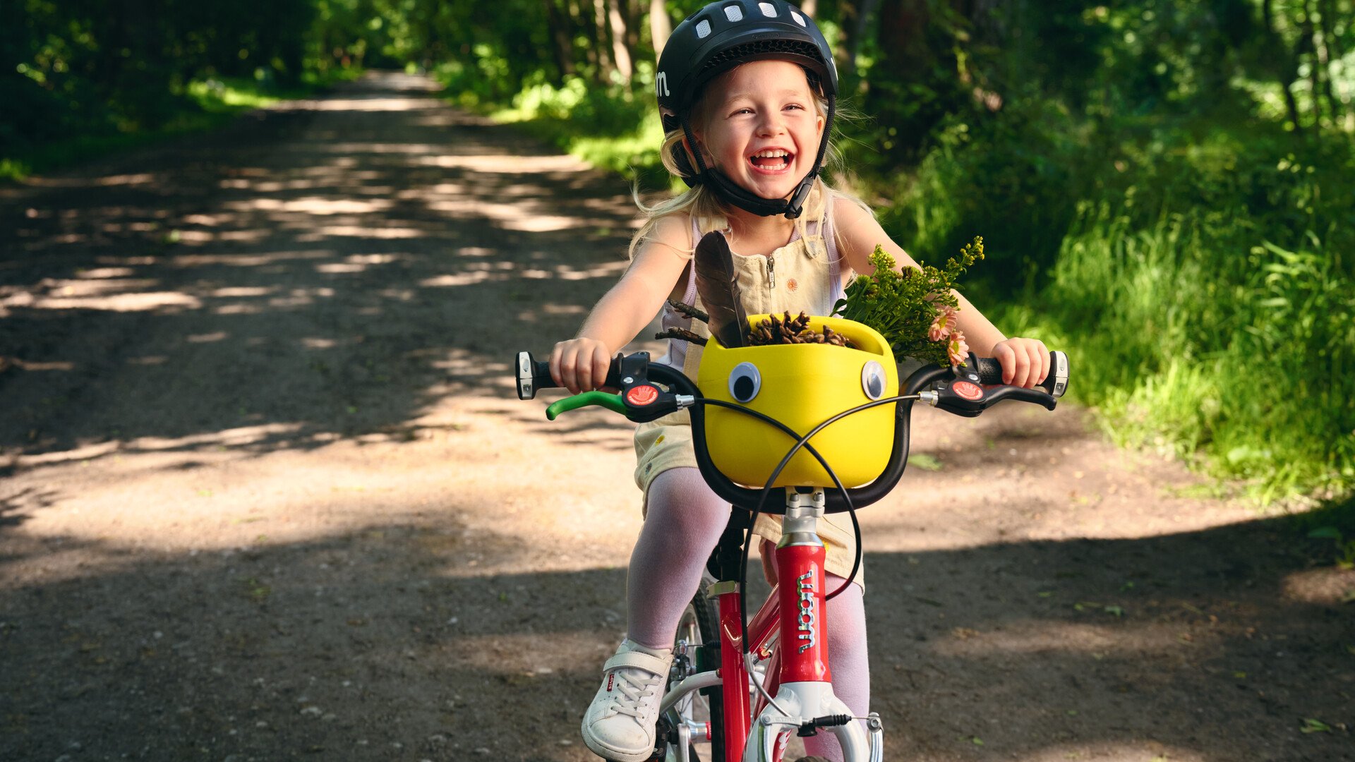 Une petite fille souriante avec un casque woom noir roule avec son vélo woom rouge et blanc sur un chemin de forêt ombragé. Sur son guidon est accroché un panier à vélo jaune rempli de petits trésors glanés dans la nature.