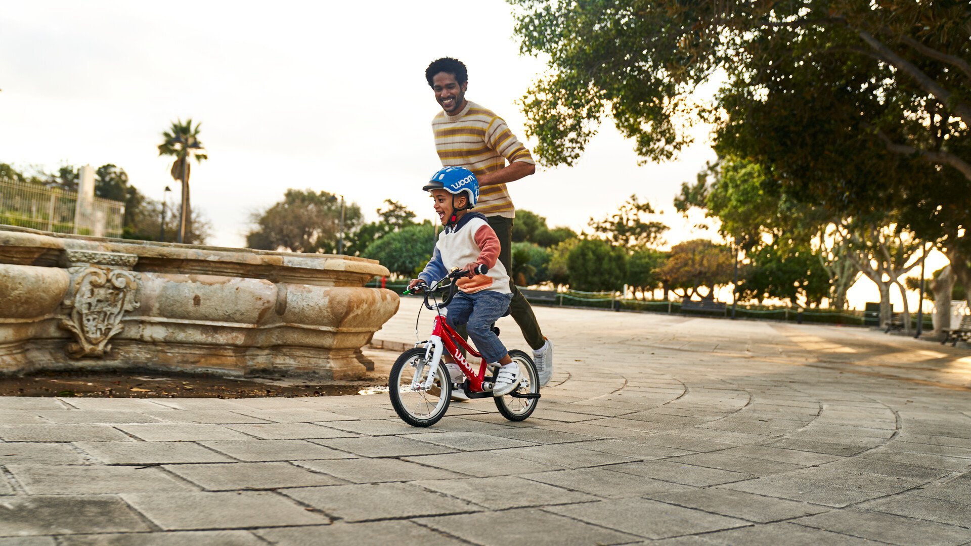 En dreng cykler på en rød woom-børnecykel, mens hans far løber ved siden af.
