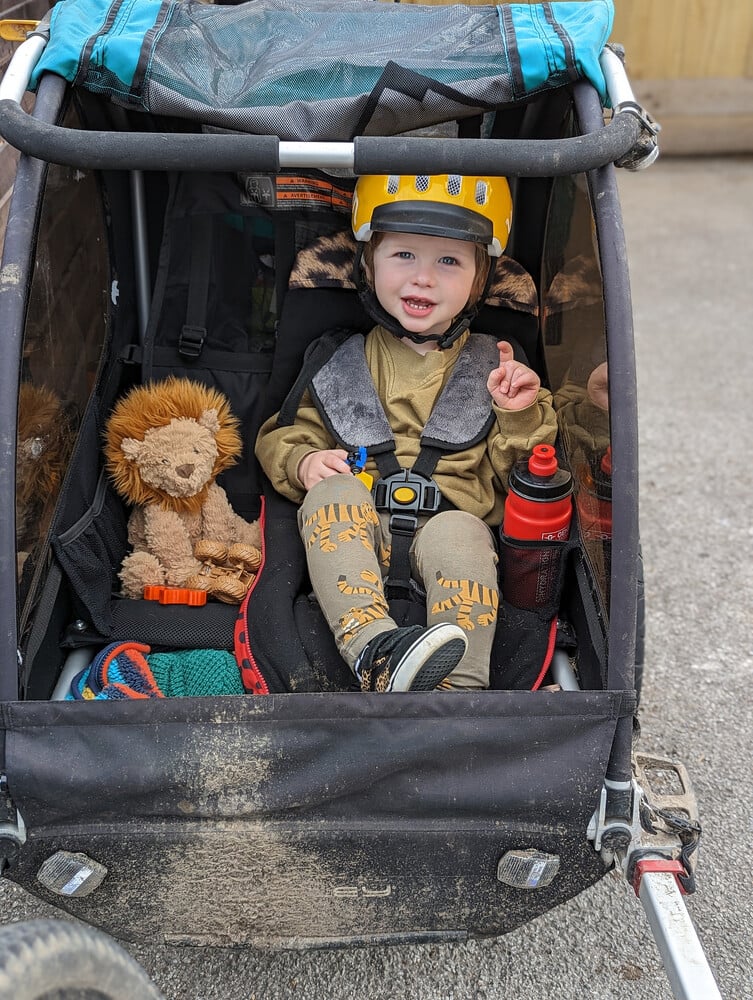 A young boy wearing a yellow woom bike helmet sits in a bike trailer with his cuddly lion by his side.