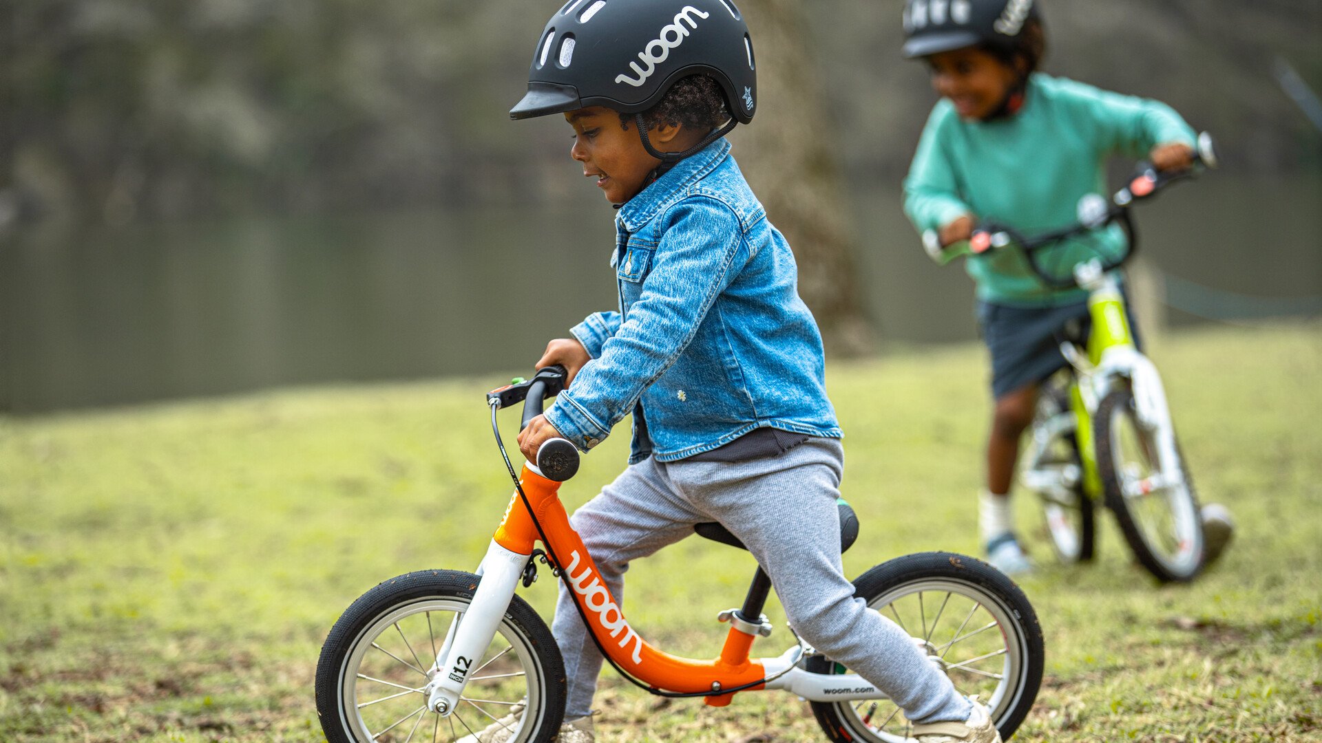 Two kids with black helmets ride orange and green woom balance bikes on some grass.