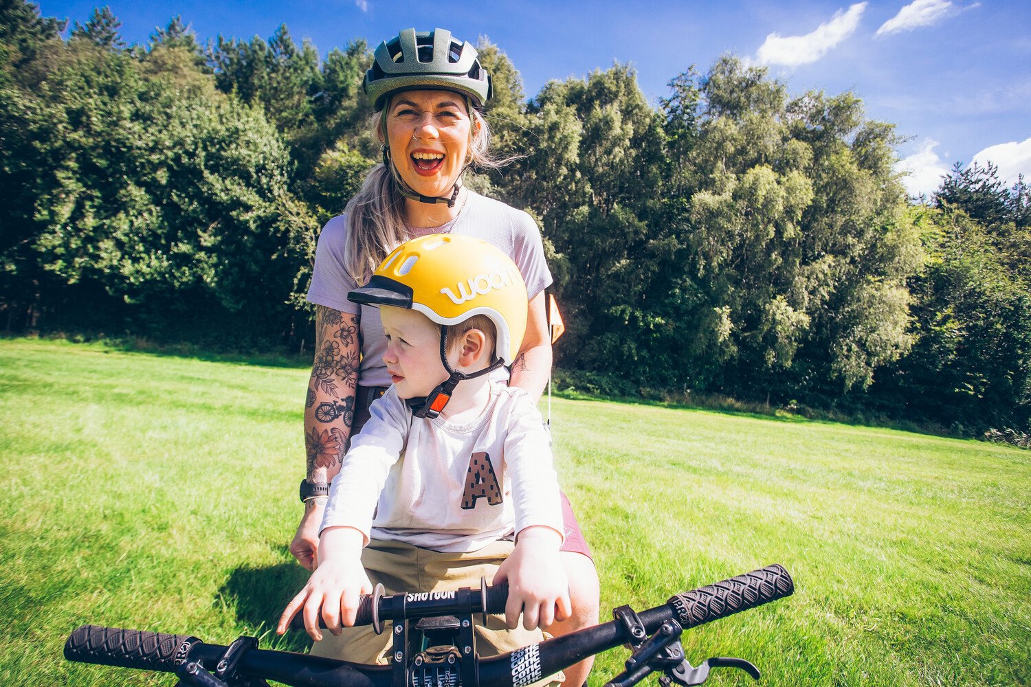 Vor einer lachenden Radfahrerin sitzt ein Junge im Kindersitz und trägt einen gelben Fahrradhelm mit weißem woom Logo.