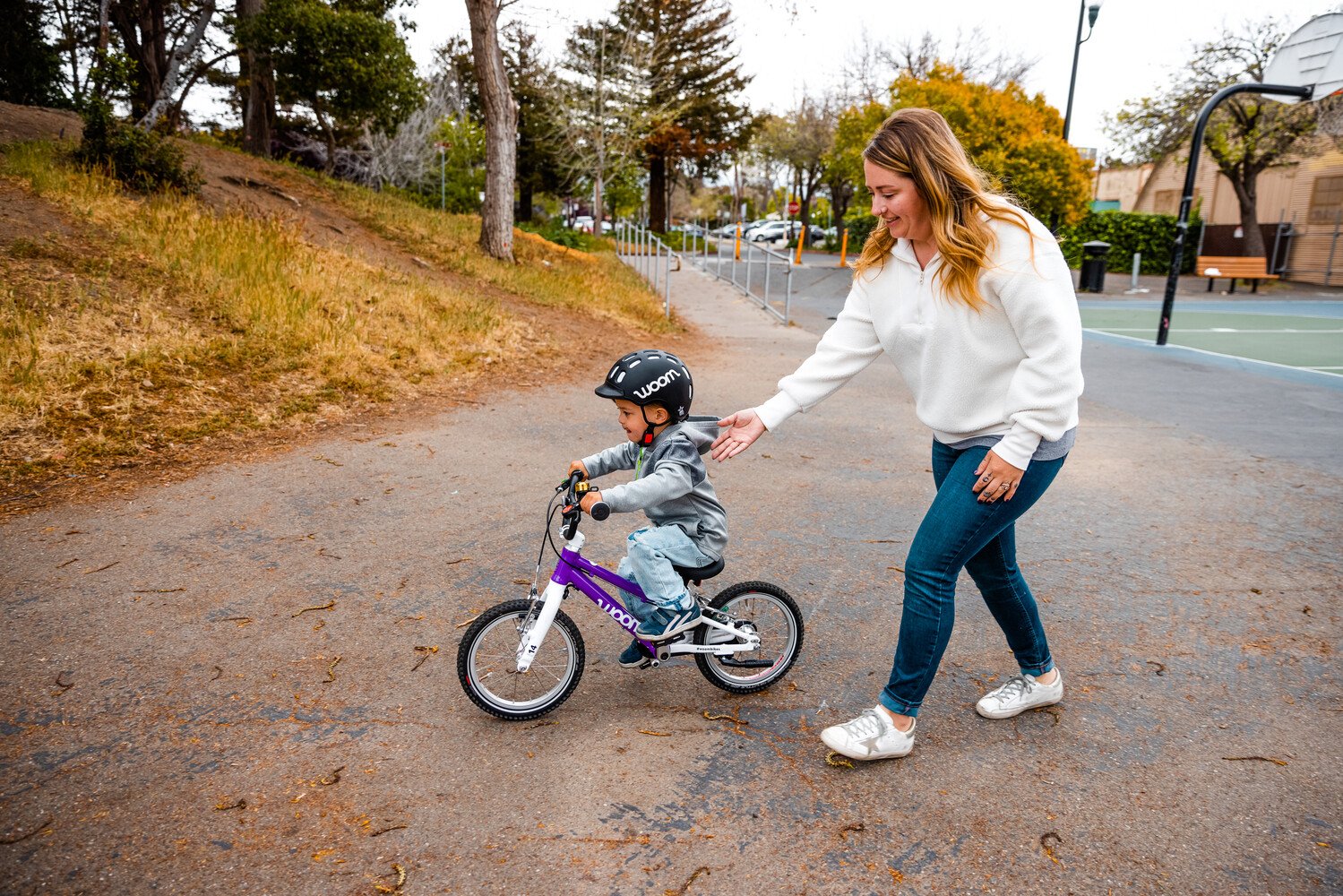 Een kind met een zwarte woom helm fietst met lichte ondersteuning van zijn moeder op een paarse kinderfiets.