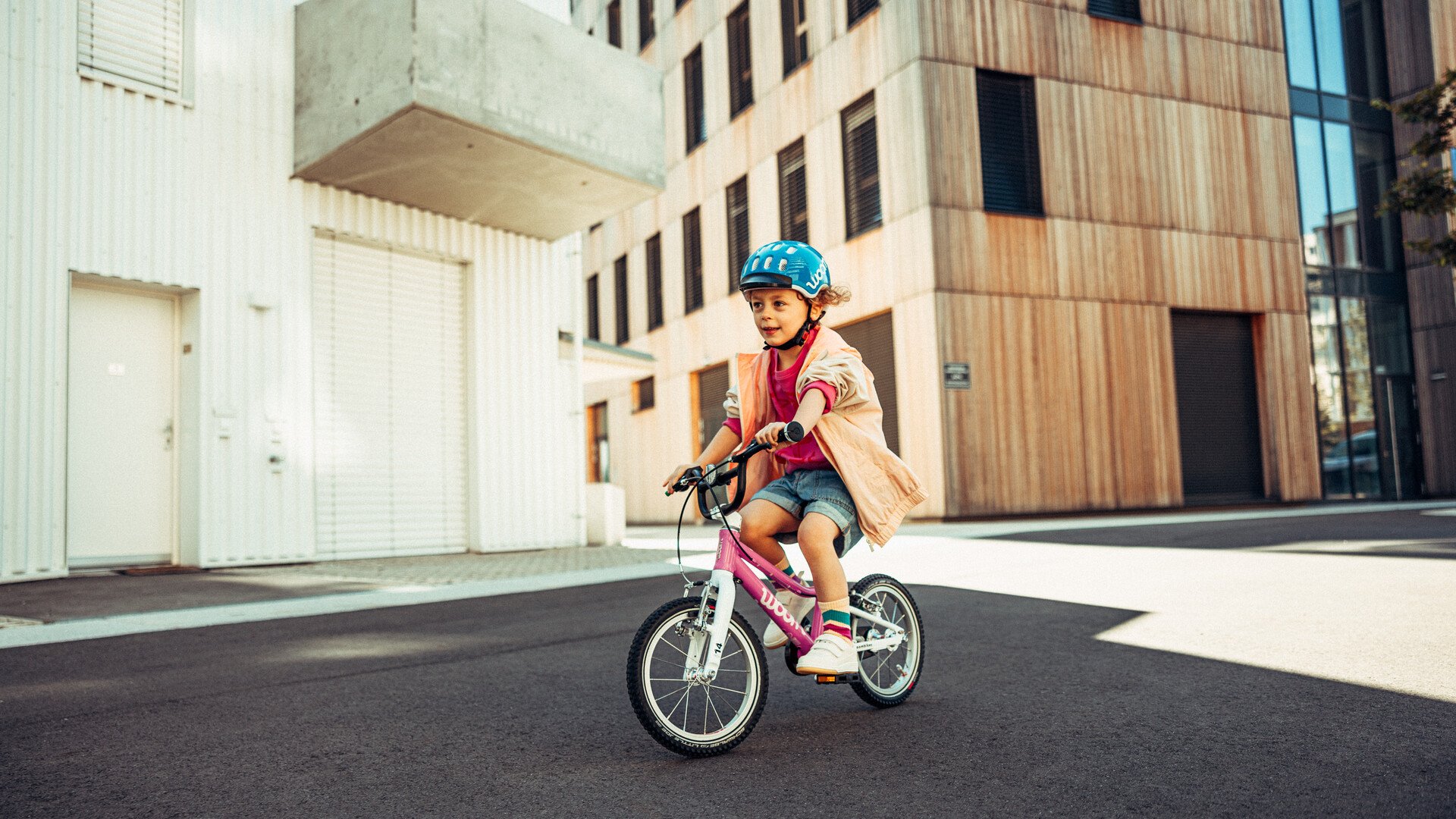 Een meisje met blauwe fietshelm fietst in een stedelijke omgeving op een roze kinderfiets van woom.