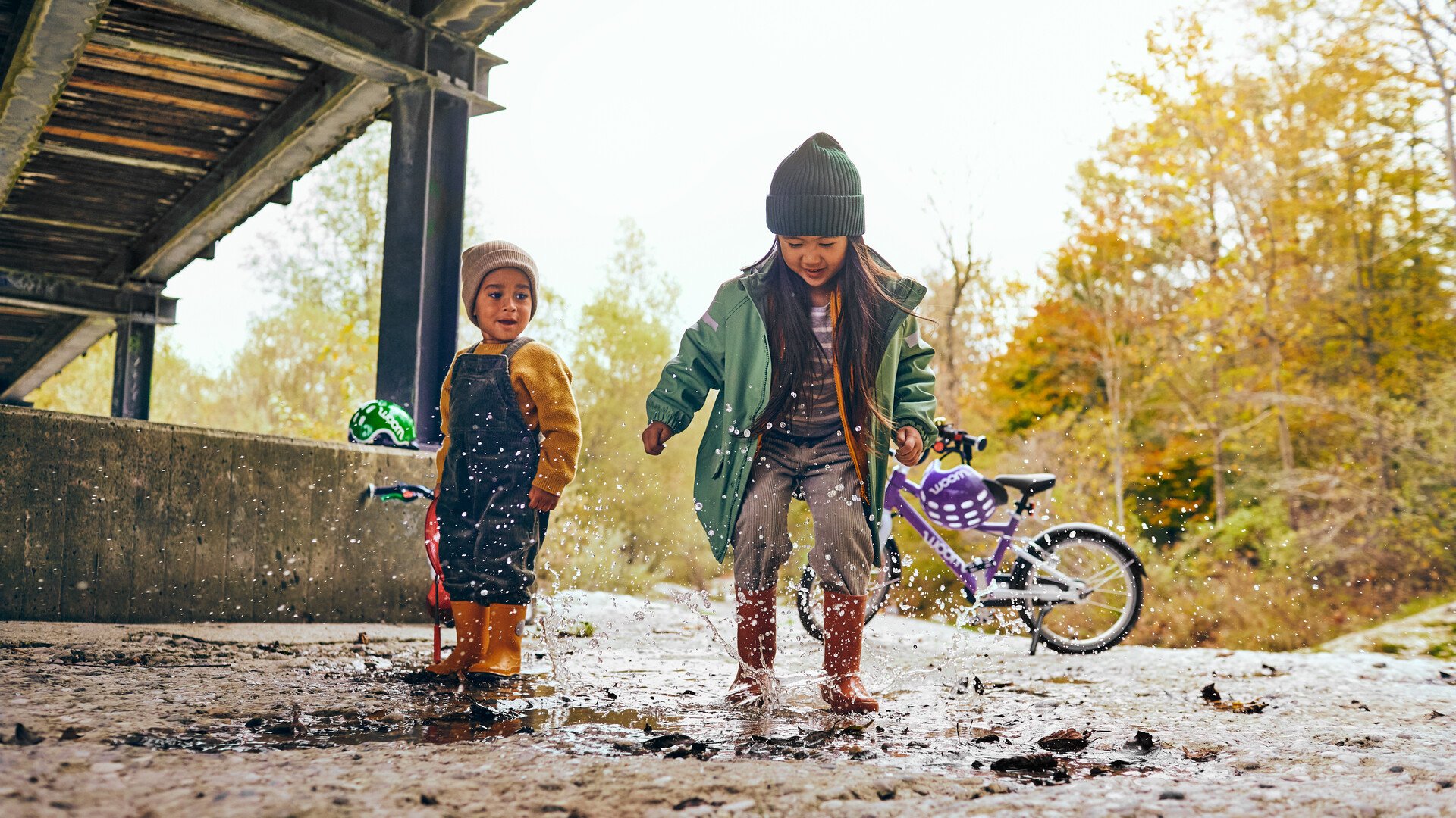 Zwei Kinder toben in einer Pfütze, Wasser spritzt hoch, im Hintergrund steht ein woom Fahrrad mit Fahrradhelm am Lenker.