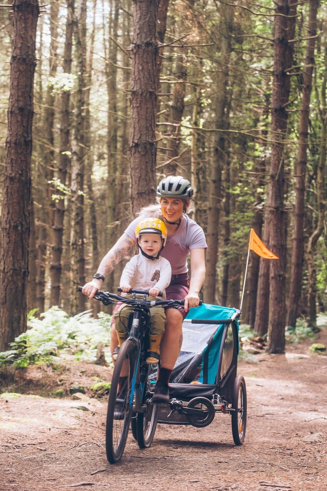 Eine Mutter radelt mit ihrem Sohn im Kindersitz – er trägt einen gelben KIDS Helm – im Wald und zieht einen Fahrradanhänger.