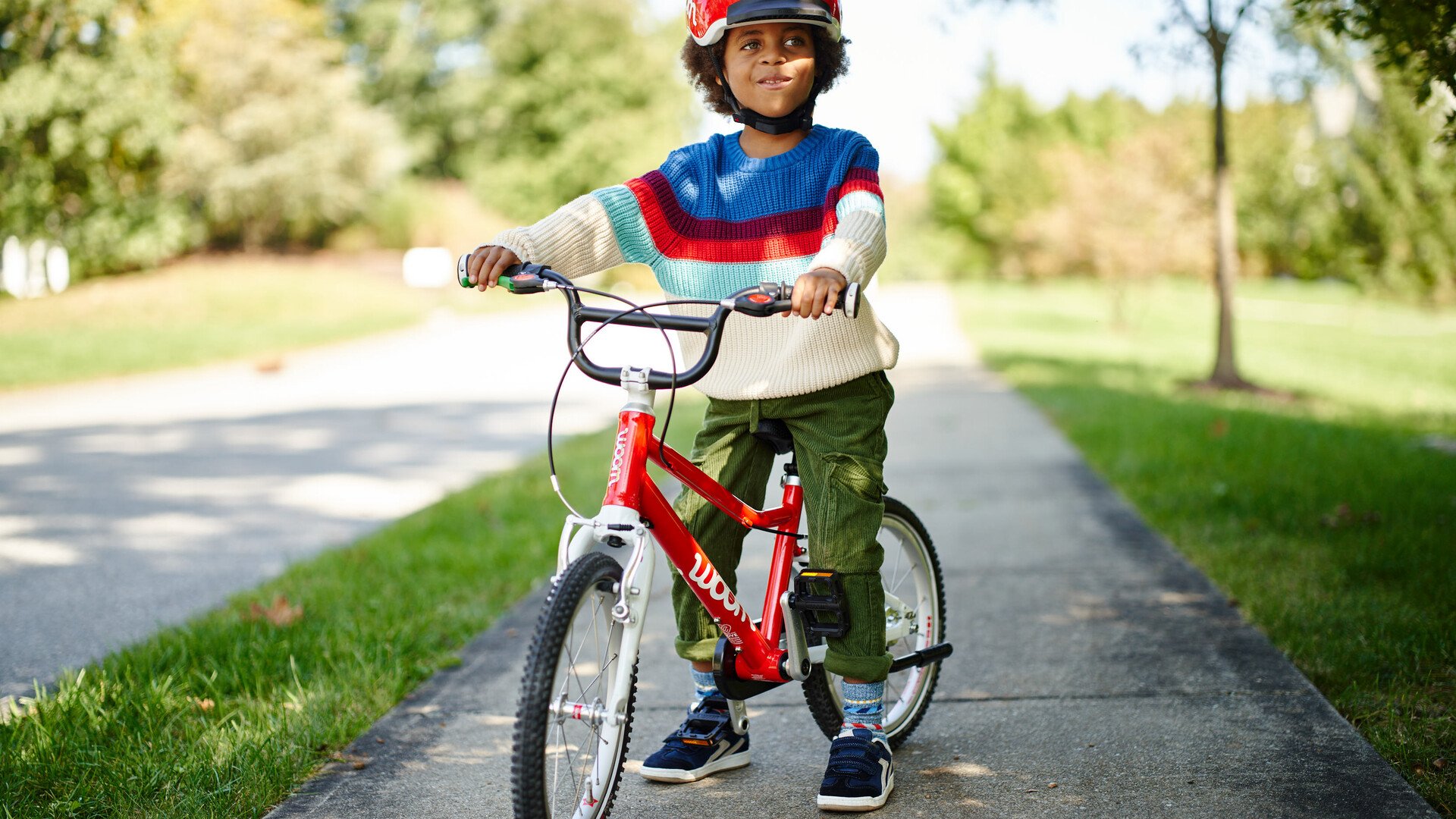Ein kleines Kind mit einem roten Helm sitzt auf einem roten woom Fahrrad und hat beide Füße auf dem Boden abgestellt.