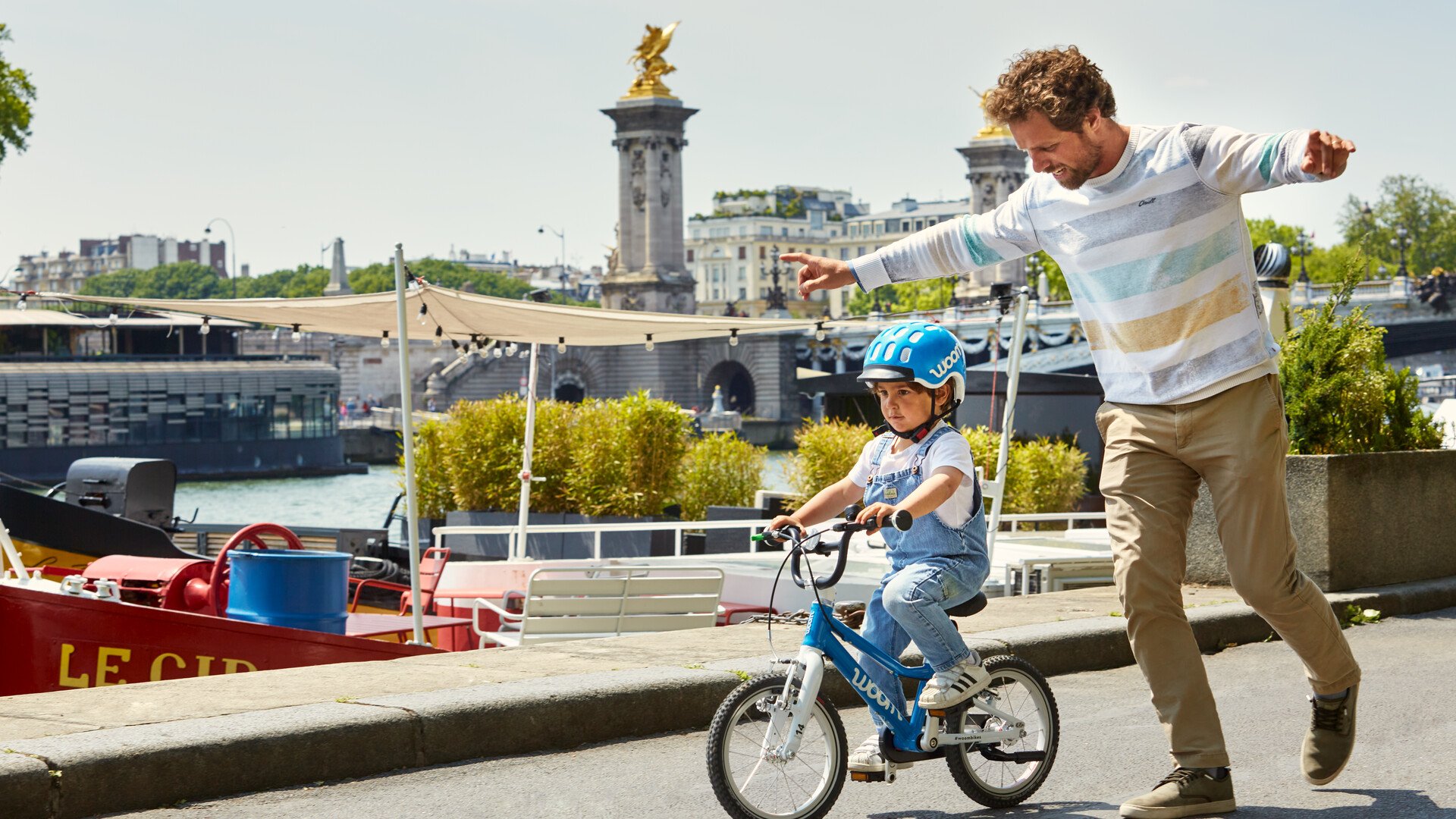 Et barn med en blå hjelm cykler koncentreret på en blå cykel, mens hans far løber ved siden af.