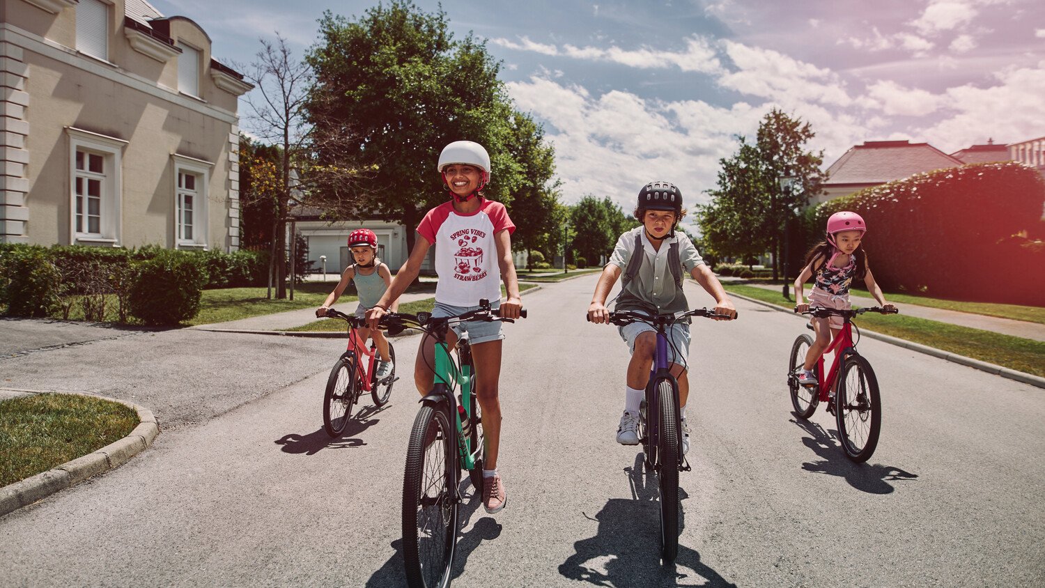 Wide surburban road with four children riding alongside each other on their woom EXPLORE bikes towards the camera.