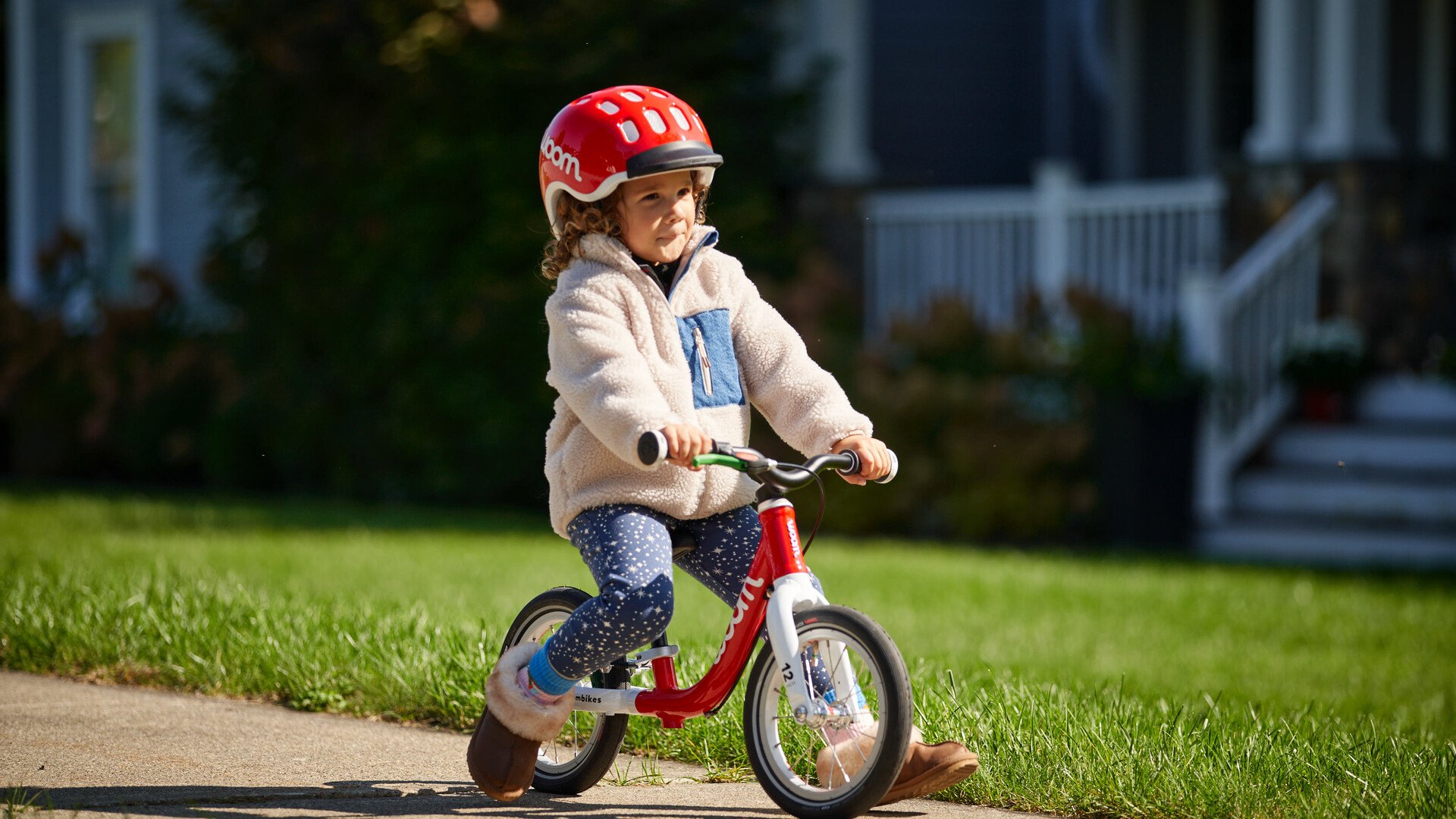 Ett lyckligt småbarn med röd hjälm och röd balanscykel av märket woom kommer körande på trottoaren.