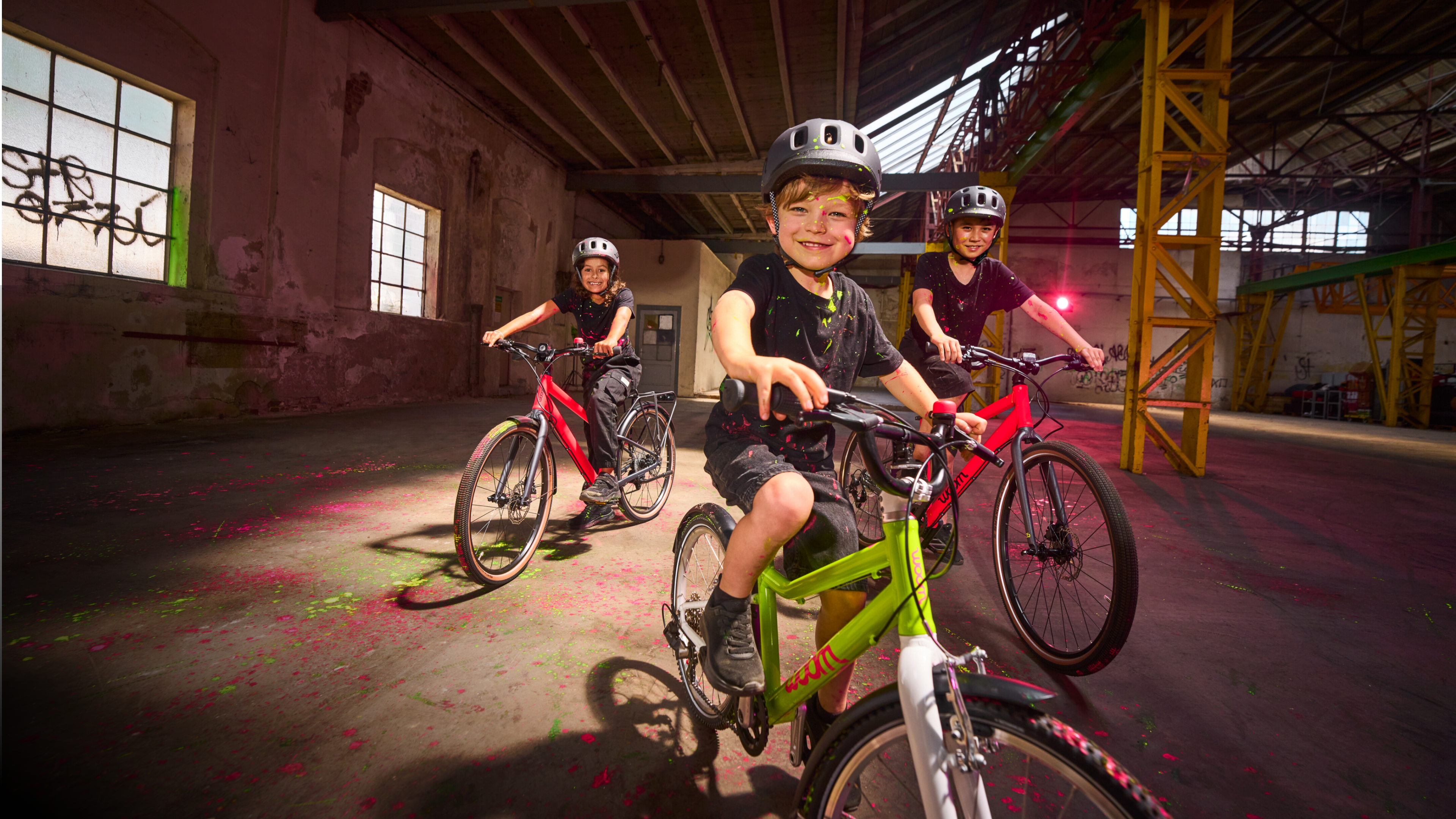 Drei Kinder mit Helmen auf neonfarbenen woom Bikes in einer alten Industriehalle mit bunten Farbspritzern.