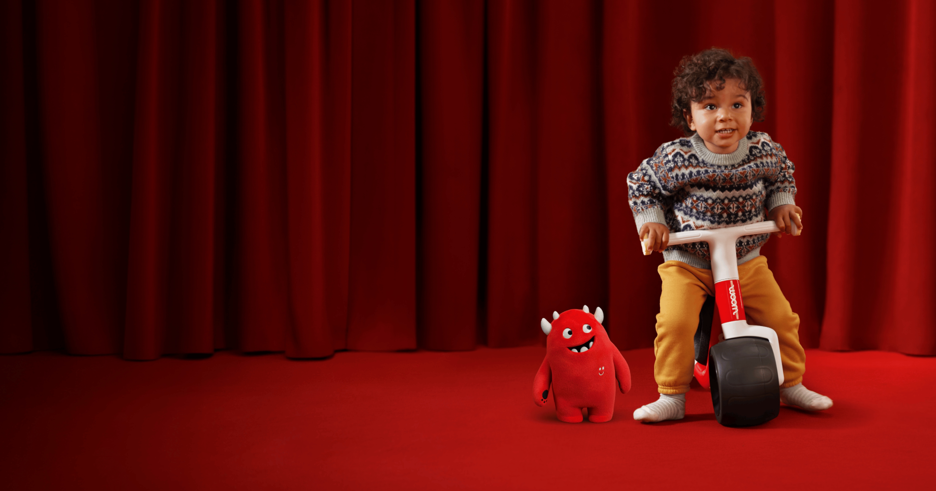 A young boy sits facing the camera atop a new red woom WOW in front of a red velvet curtain and on top of a red carpet. Buddy, the red woom monster mascot, stands to the left of the bike smiling up at the boy.