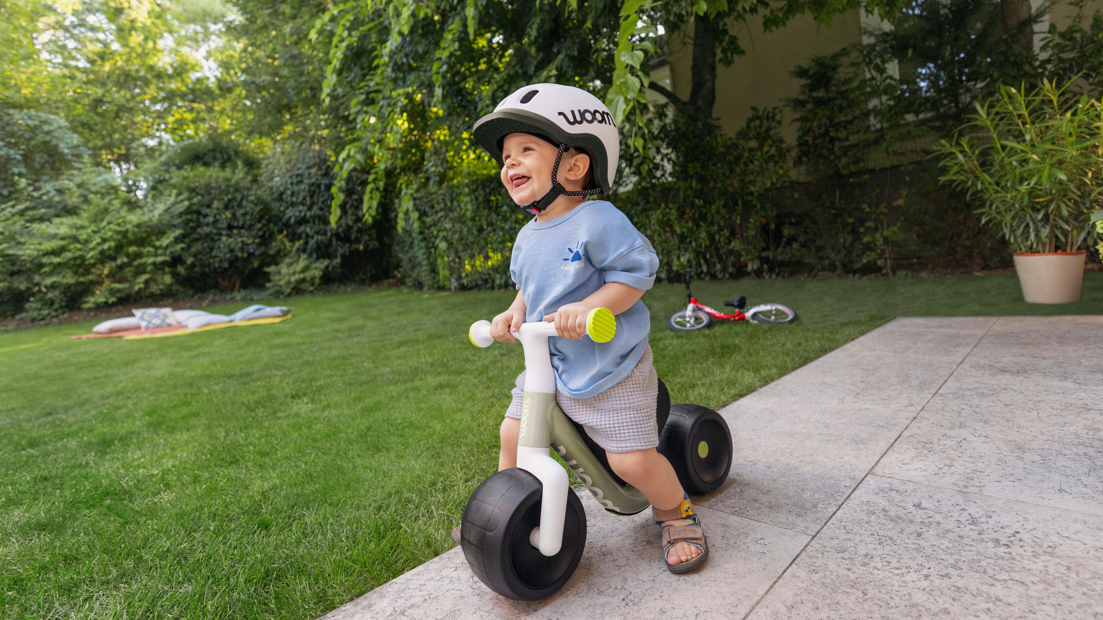 Un enfant souriant portant un casque avec sa draisienne dans un jardin. Bannière publicitaire pour les woom WOW et woom GO.
