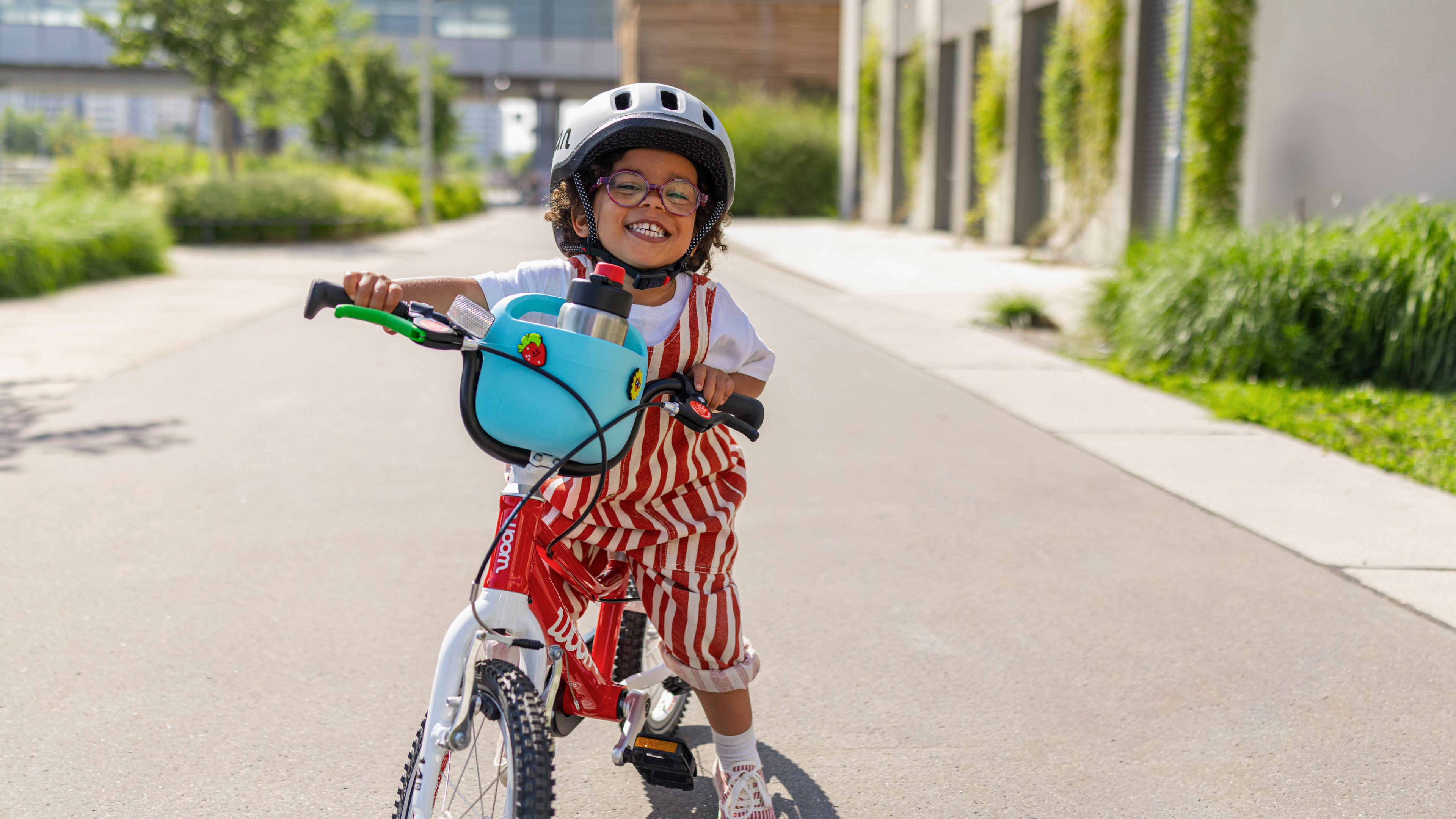 Un enfant souriant avec un casque enfant READY roule sur un vélo woom GO rouge équipé d’un panier POP bleu par temps ensoleillé.