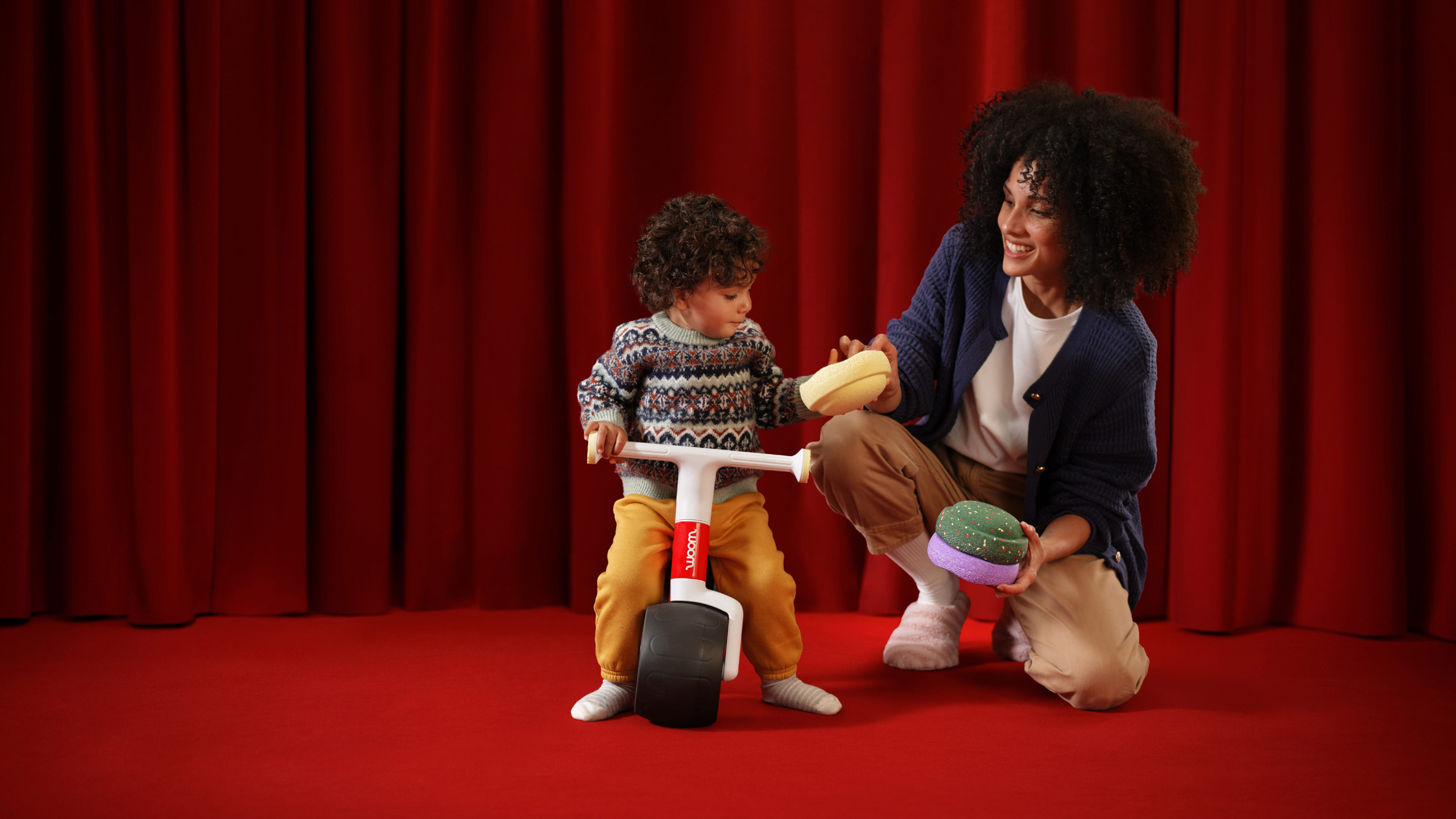 A toddler sitting on the red woom WOW Seasonal Colour Edition balance bike on a red stage, while a woman cheers in the background near scattered exclusive Stapelstein® Original Minis.