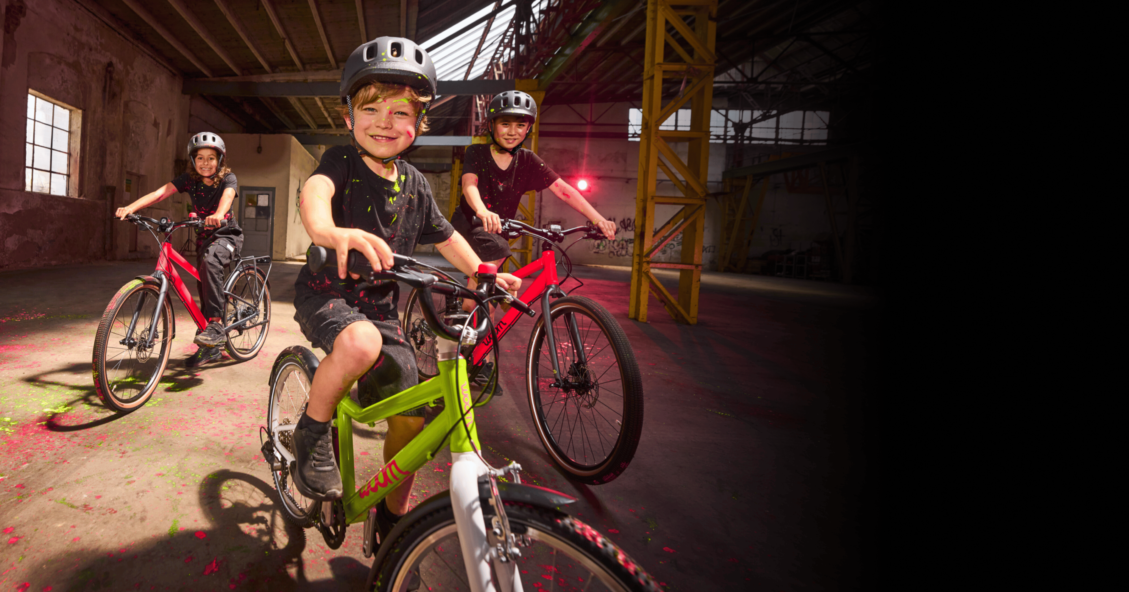 A photo of three kids riding their bikes through a paint-splattered warehouse, highlighting the electric new colors featured on each bike: neon lime woom GO and neon pink woom EXPLORE.