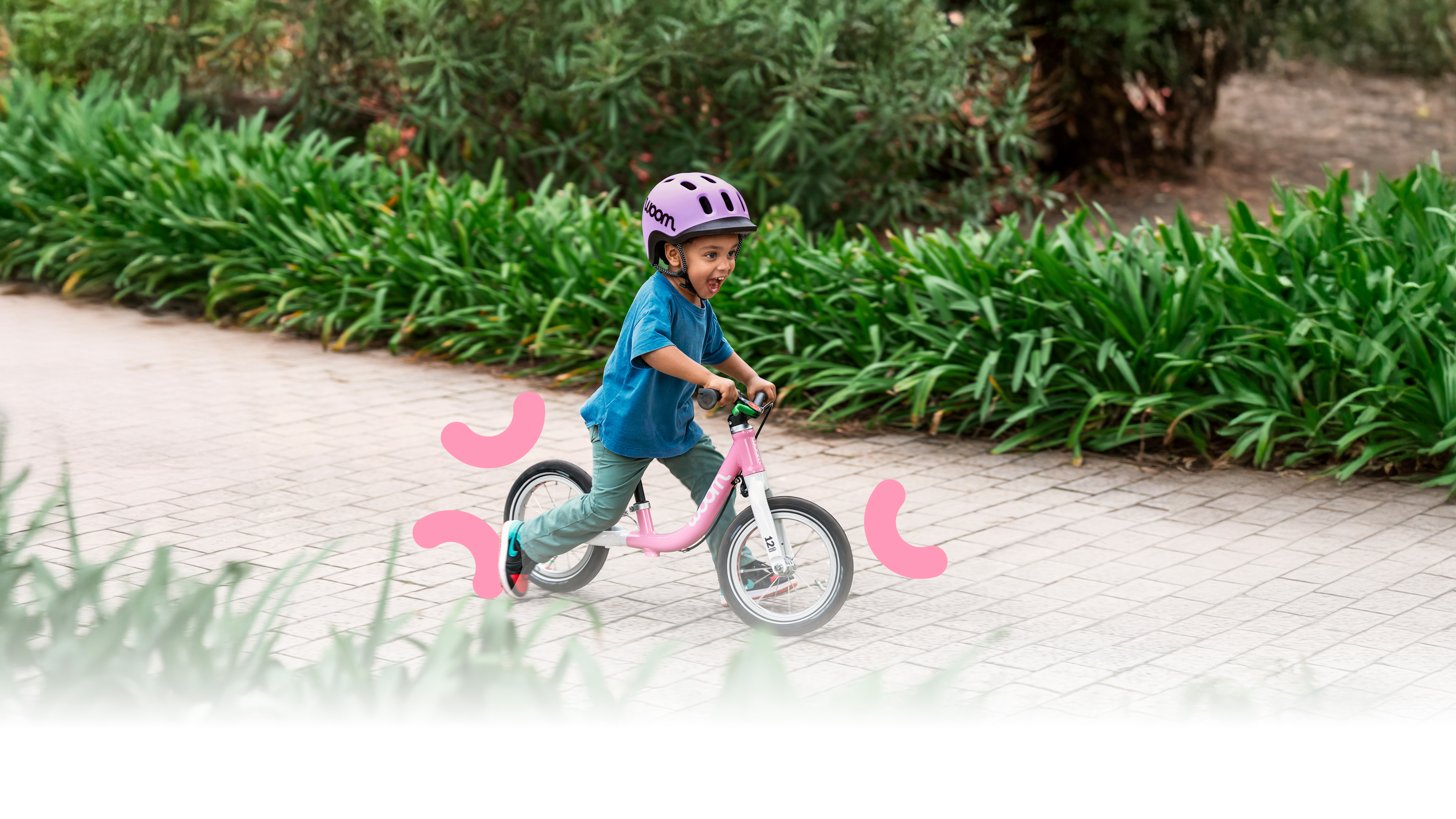 Un enfant portant un casque READY violet roule avec sa woom GO rose sur un chemin pavé dans un cadre vert et fleuri.