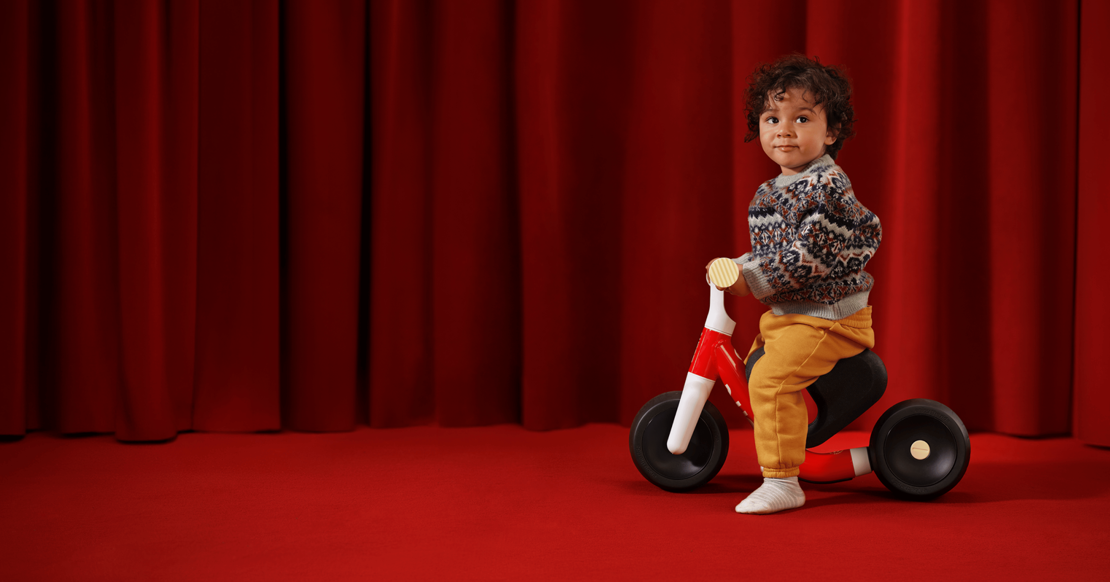 A young boy sits in profile atop a new red woom WOW in front of a red velvet curtain and on top of a red carpet.