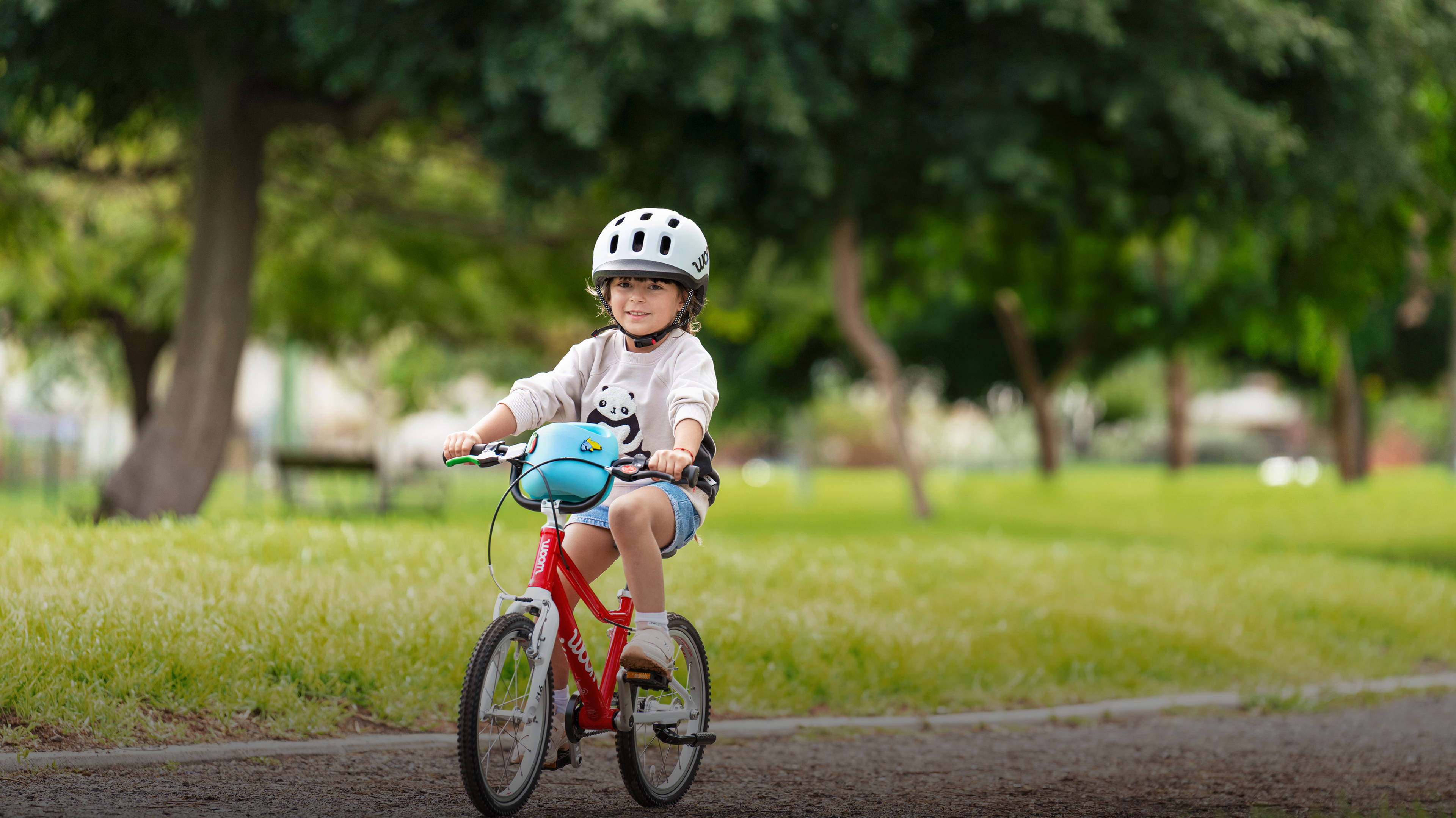 Kind mit woom READY Helm fährt auf einem roten woom GO, während eine Frau im Park zuschaut.