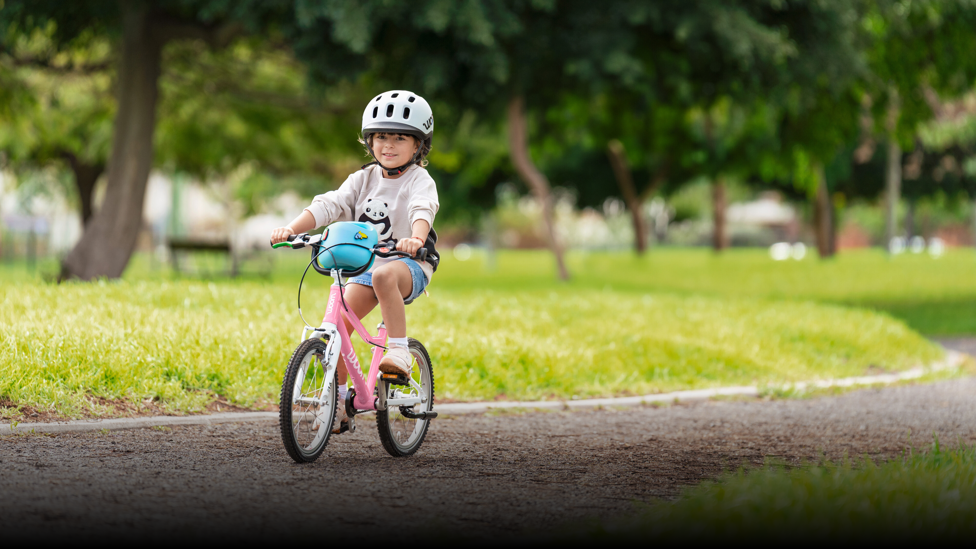 Un enfant avec un casque READY gris pédale sur un vélo woom GO powder pink ; une femme l’encourage, les bras ouverts. 