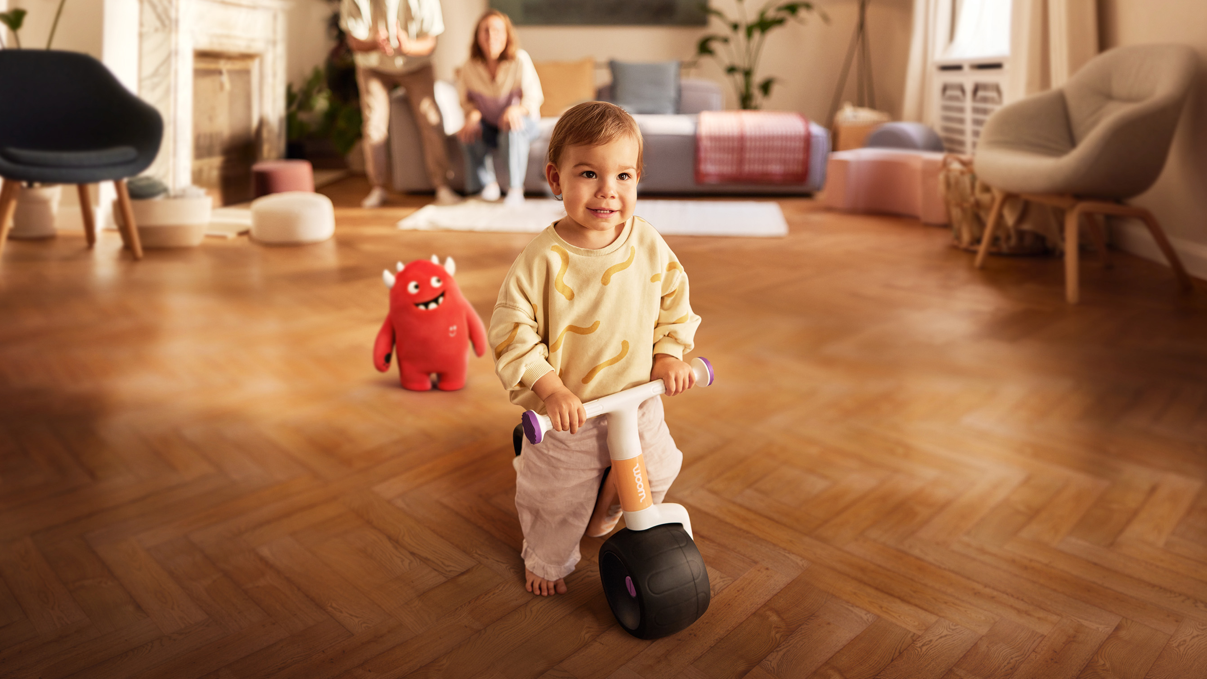 Child sitting on woom WOW balance bike, smiling, parents in the background.