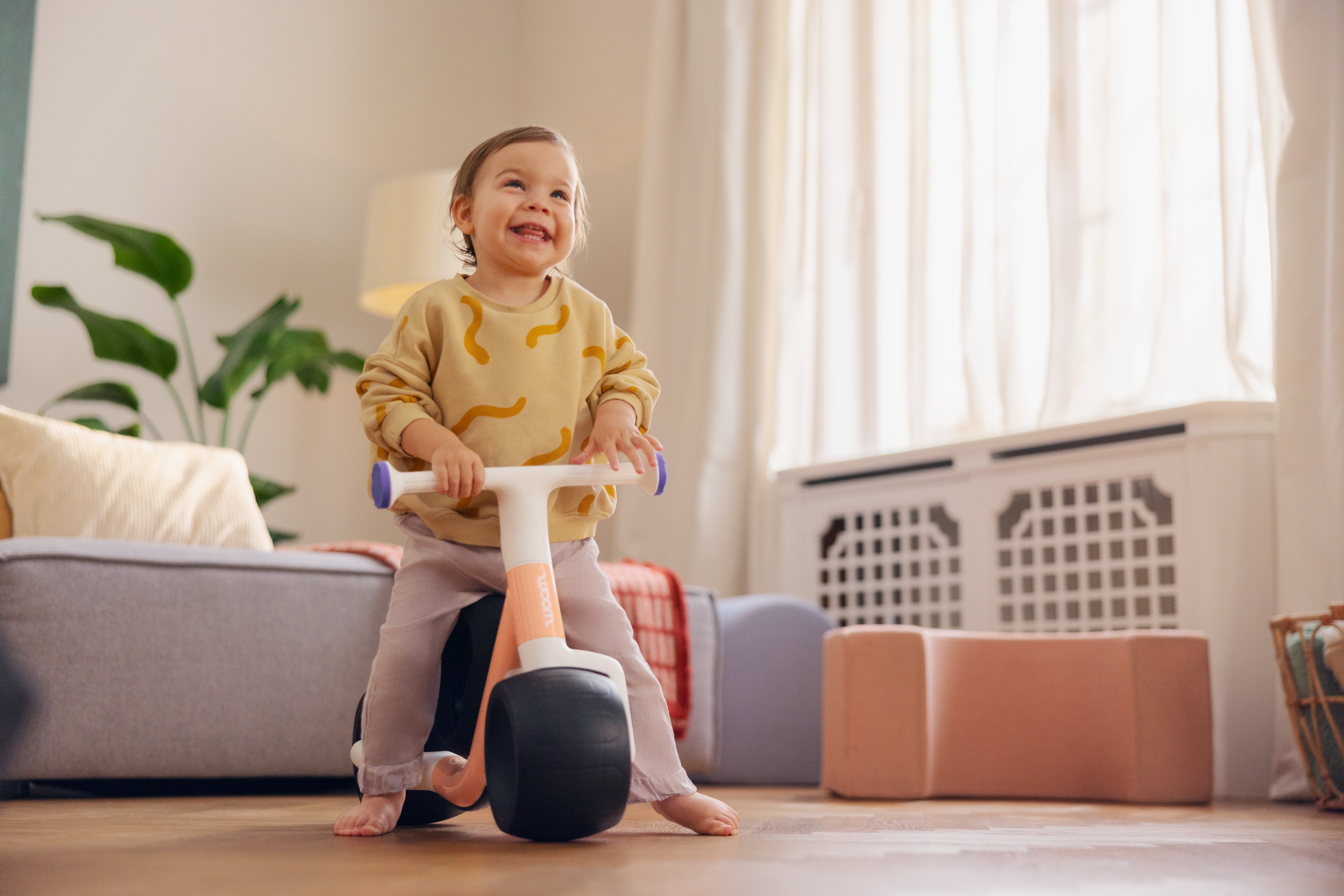 A smiling young child hold the white handlebars on an orange-colored woom WOW balance bike with black wheels.