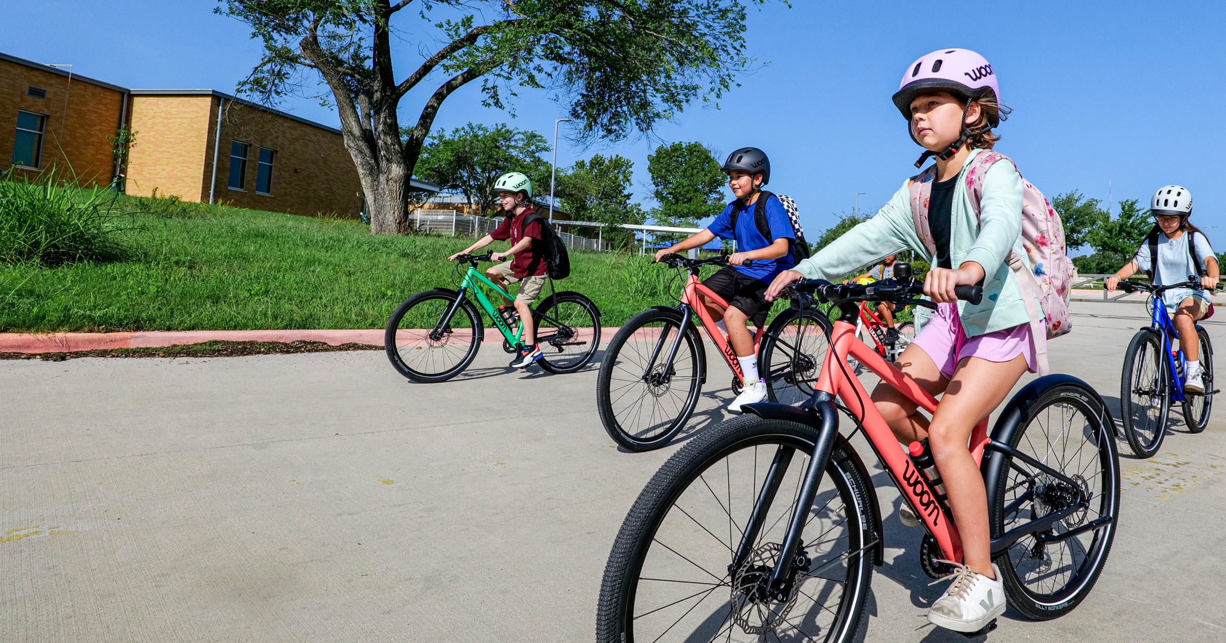 A group of young bike riders steer their woom EXPLORE bikes through a school parking lot. They are wearing woom READY helmets and their magnetic blue and neon coral woom EXPLORE bikes stand out against a bright blue sky.