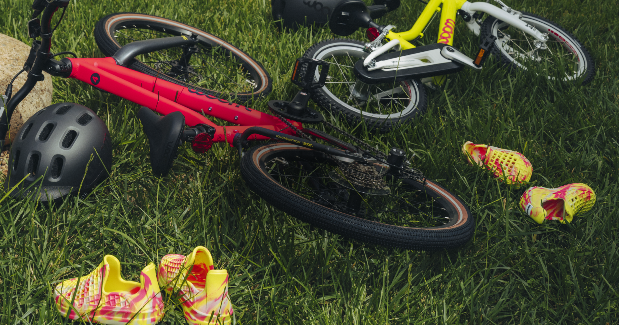 Two NEON woom bikes, a GO and an EXPLORE, lay in the grass next to a pair of neon swirl Lusso Cloud shoes.