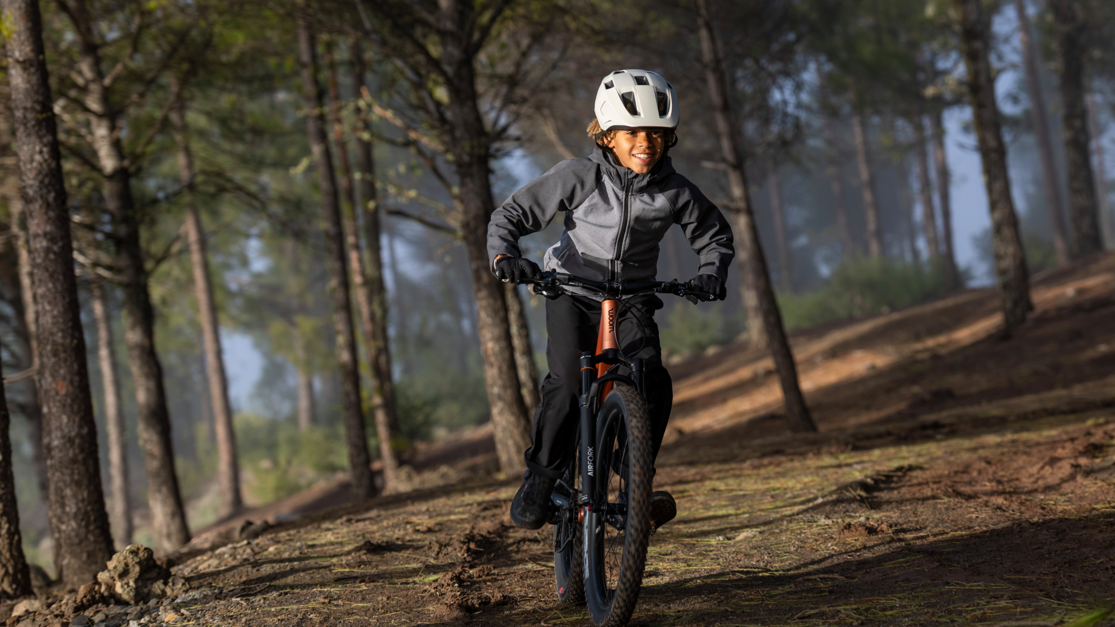 Smiling boy riding a woom OFF mountain bike through a sunny forest.