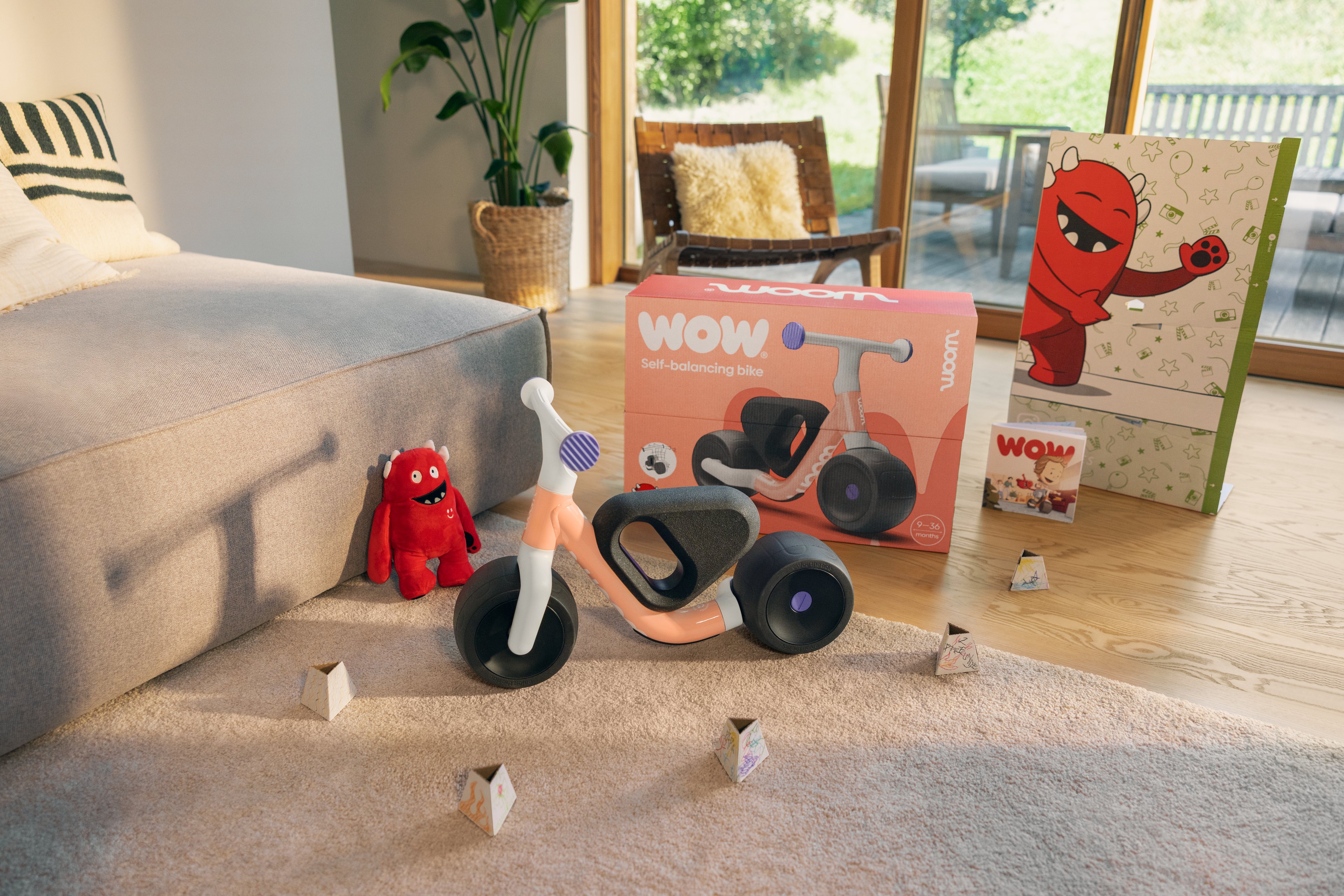 Een woom WOW loopfiets in pop peach staat samen met de originele doos en accessoires (waaronder een rode knuffel en een poster) uitgestald in een lichte woonkamer naast een zitzak.