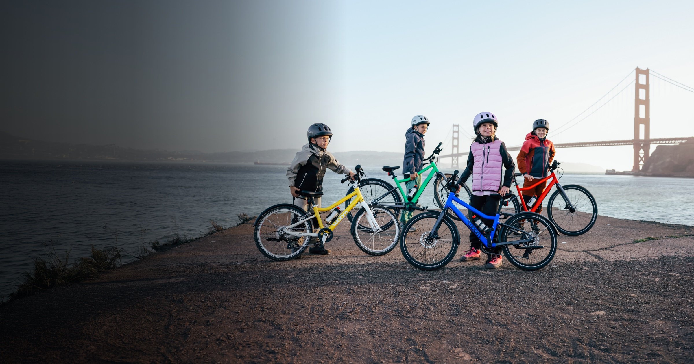Kids pose with their colorful woom GO and EXPLORE bikes in front of the Golden Gate Bridge on a chilly day.