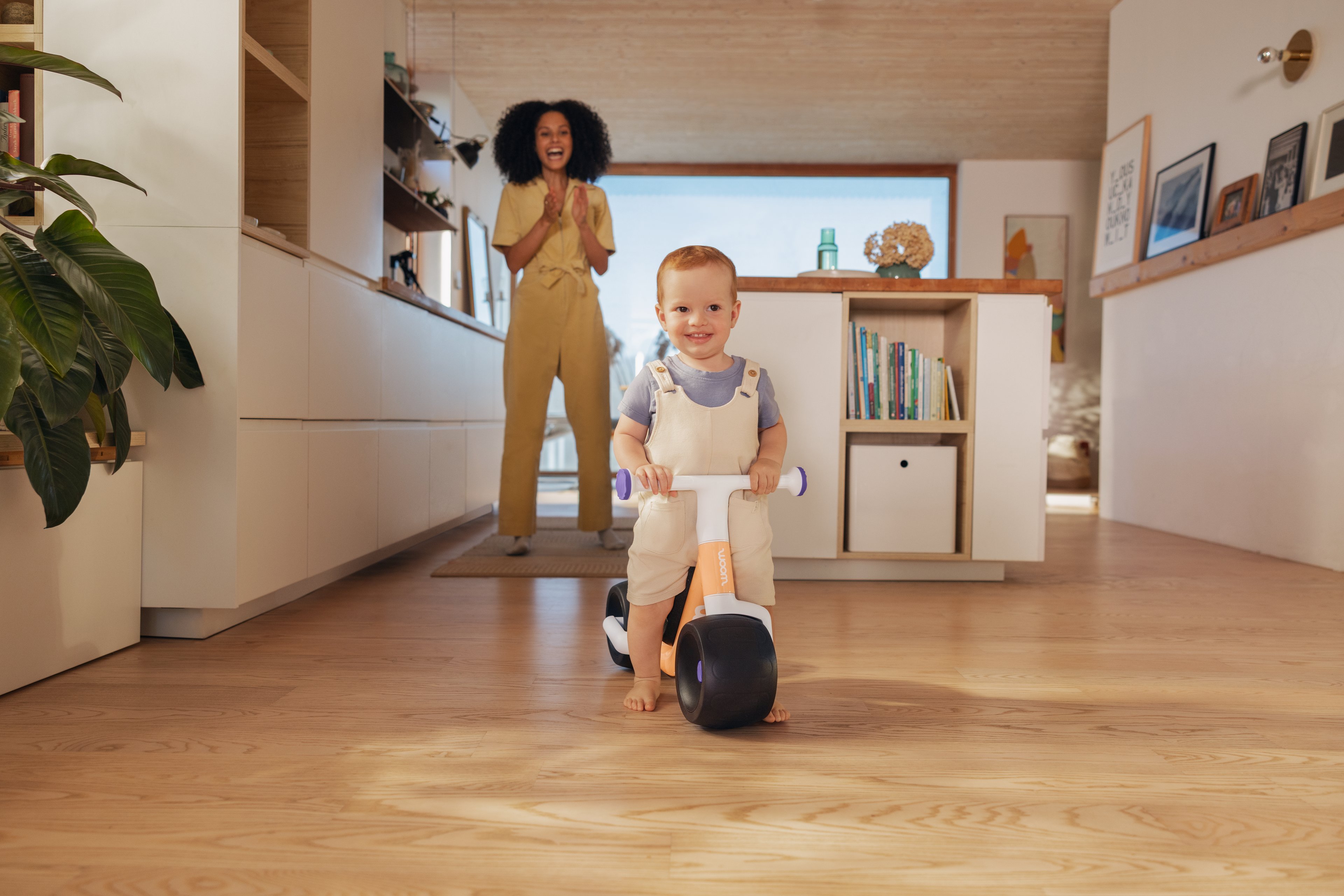 A child on an orange woom WOW balance bike smiles broadly while the mother looks on encouragingly.