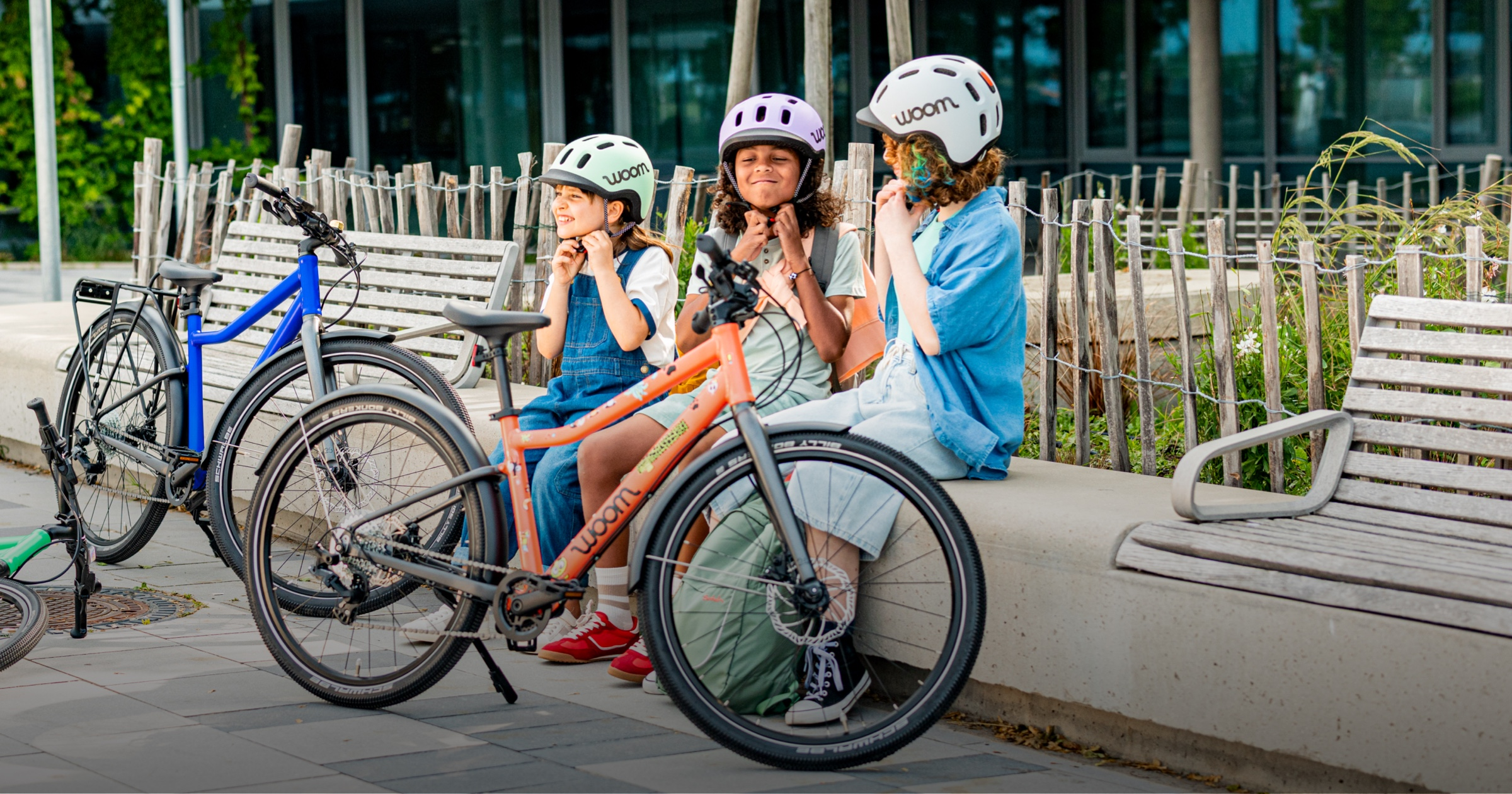 Three kids sit on a bench next to their woom EXPLORE bikes while strapping on their READY helmets.