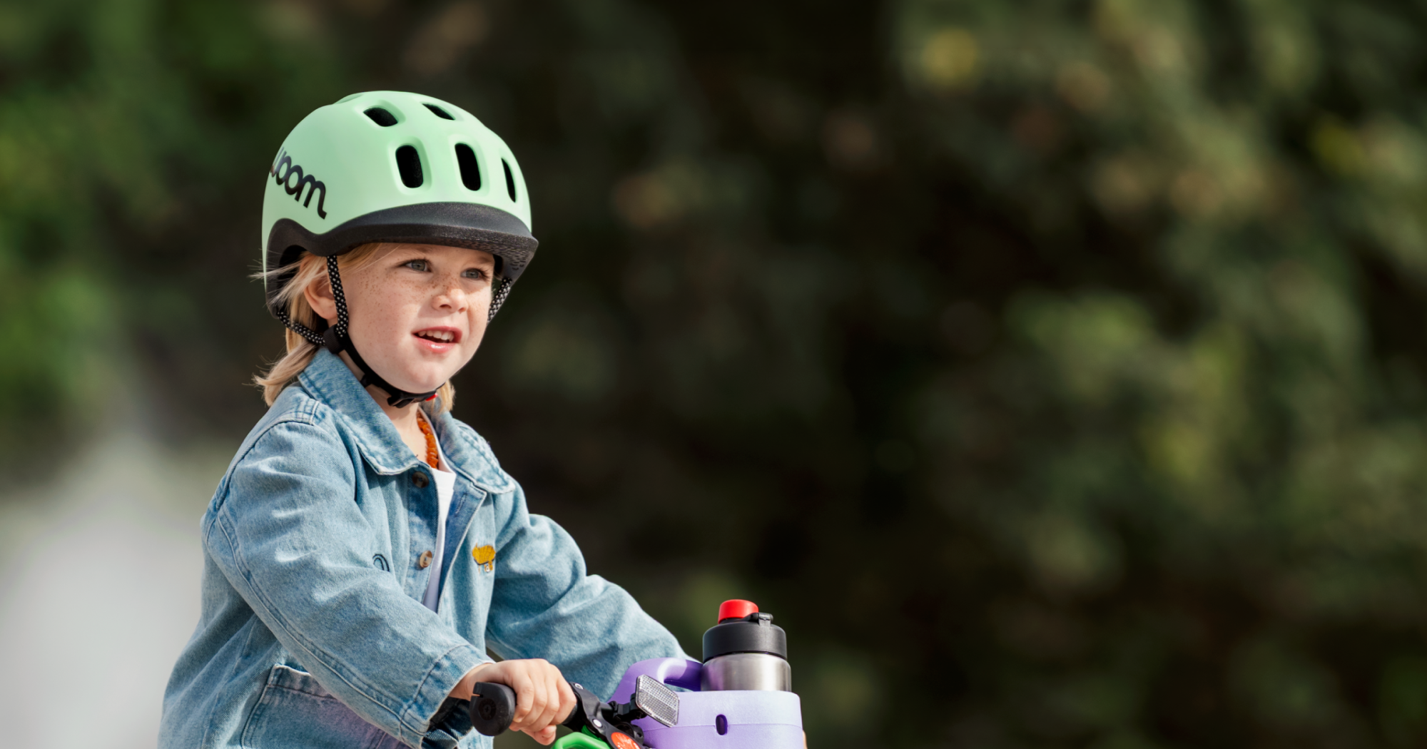 A blonde boy wears a mint READY Helmet while riding his woom GO bike with a POP Basket holding a GLUG Water Bottle attached.