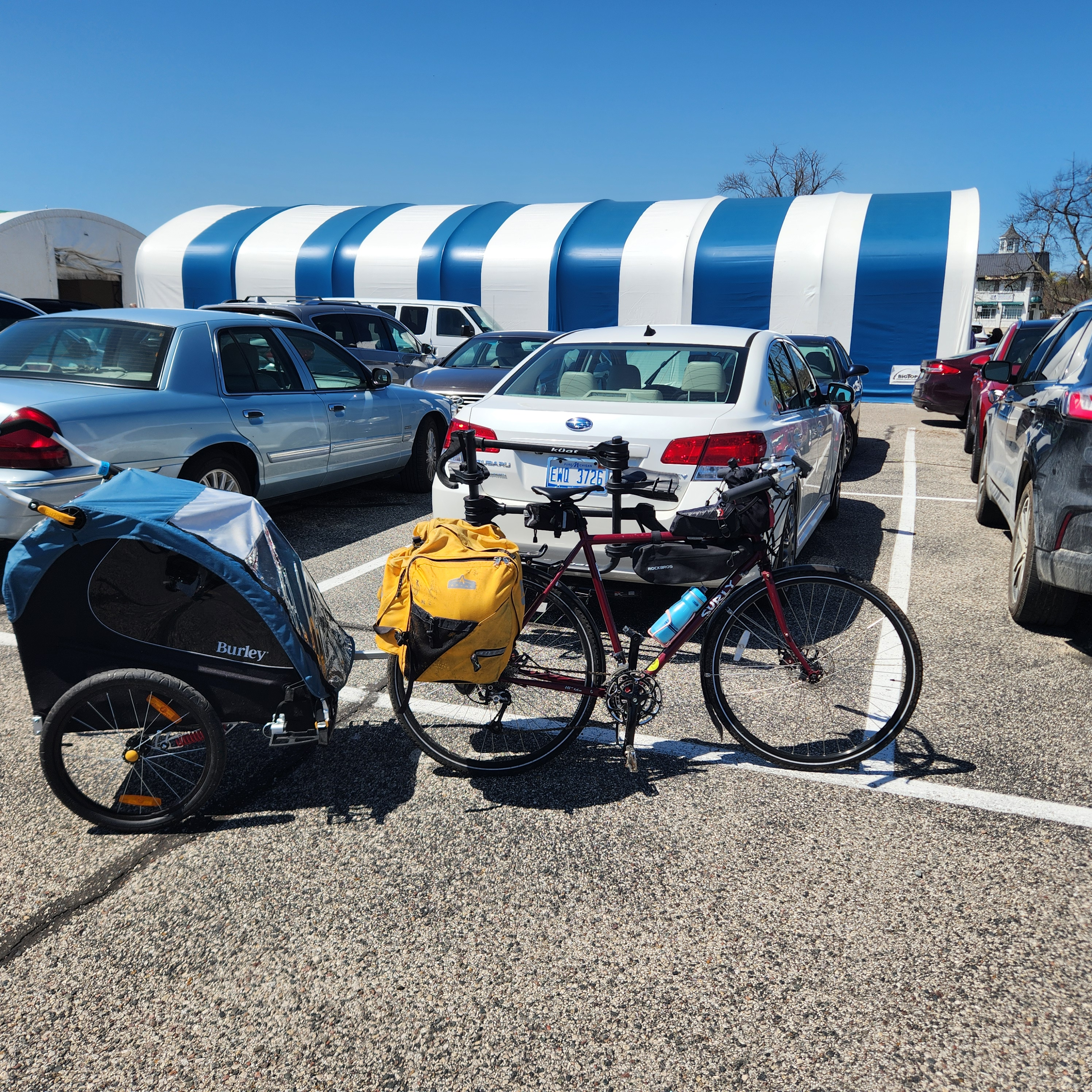 A red Surly Long Haul Trucker mens’ bicycle with water bottle, panniers, and Burley Encore bike trailer attached, standing alone near a row of cars in the parking lot of the Mackinac Island Ferry.