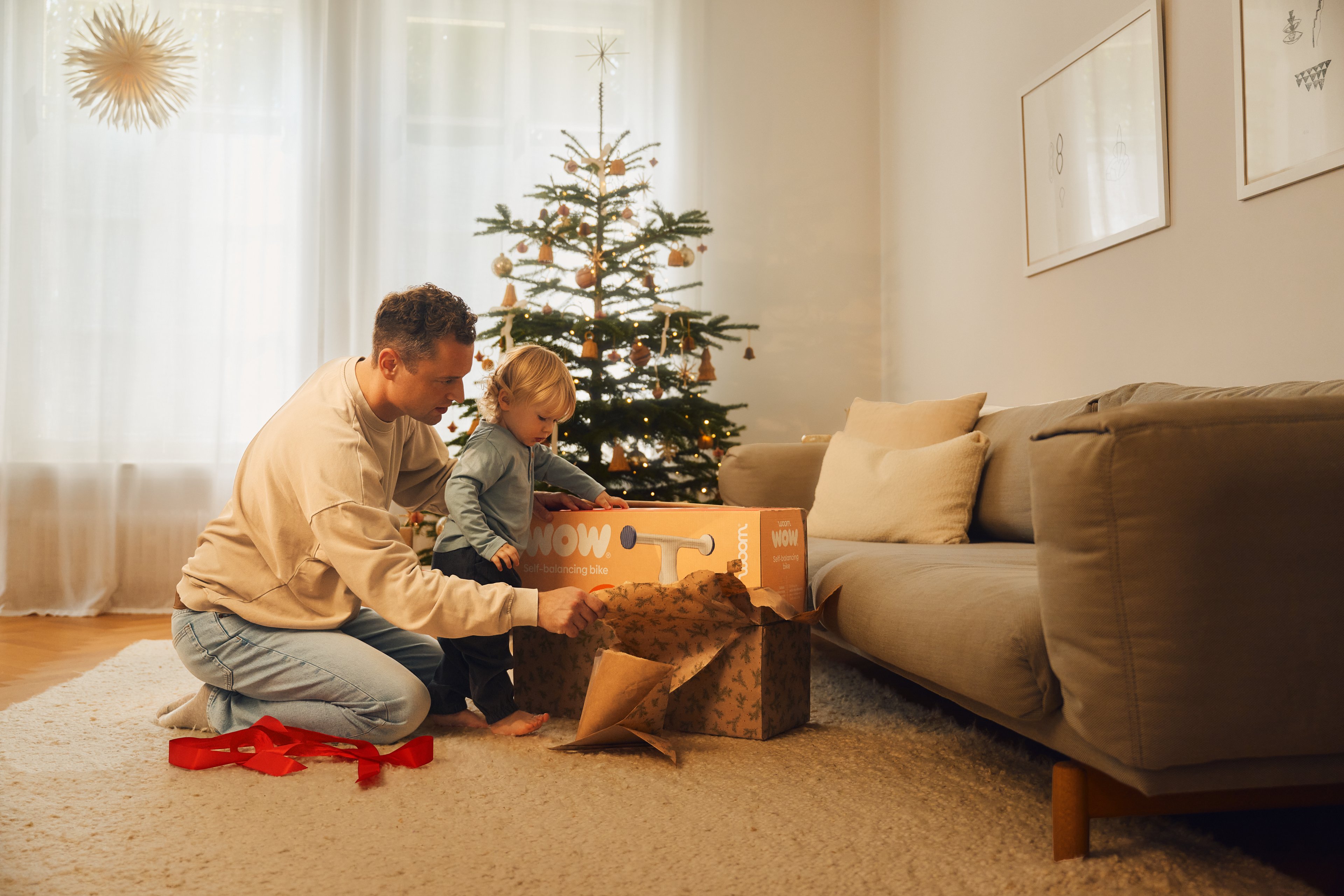 A man and a young child are unwrapping a woom WOW box in a living room with a decorated Christmas tree in the background.