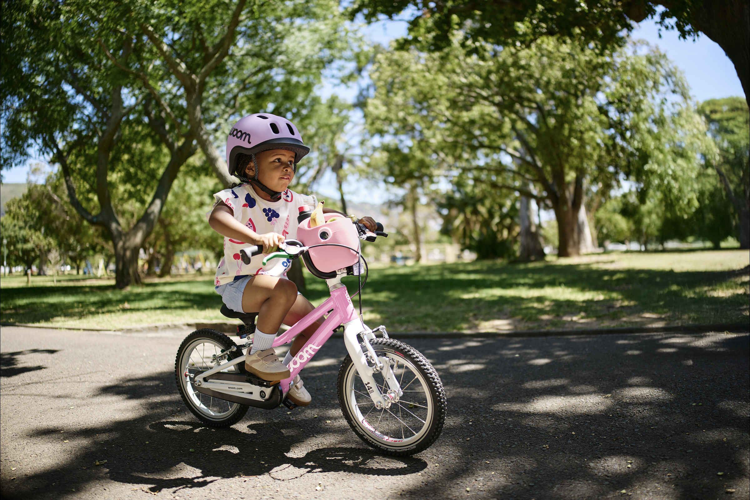 Ein Mädchen mit READY Kinderhelmen fahren auf rosafarbenen woom GO 2 Fahrrädern auf einem asphaltierten Weg im Grünen.